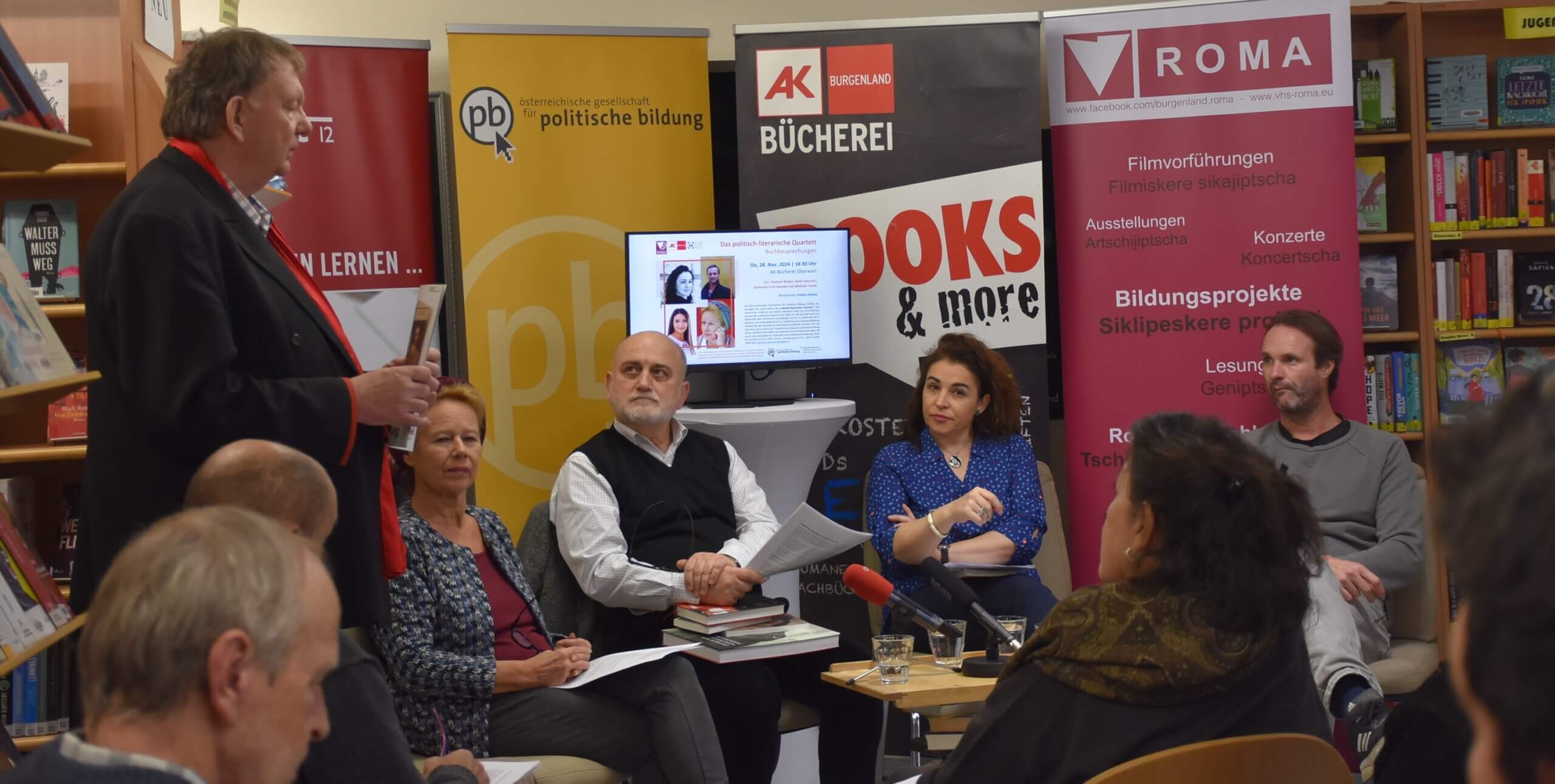 Diskussionsrunde in einer Buchhandlung mit mehreren Personen, die vor Bannern der politischen Bildung sitzen.