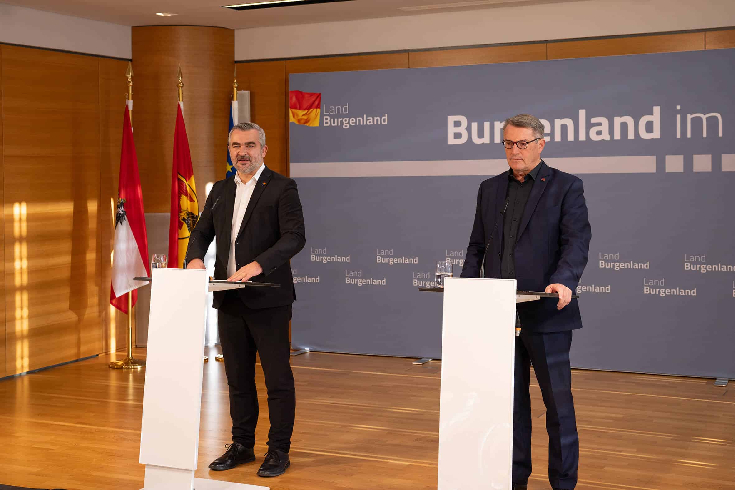 Zwei Männer halten eine Pressekonferenz im Landhaus Burgenland, neben österreichischen und Landesflaggen.