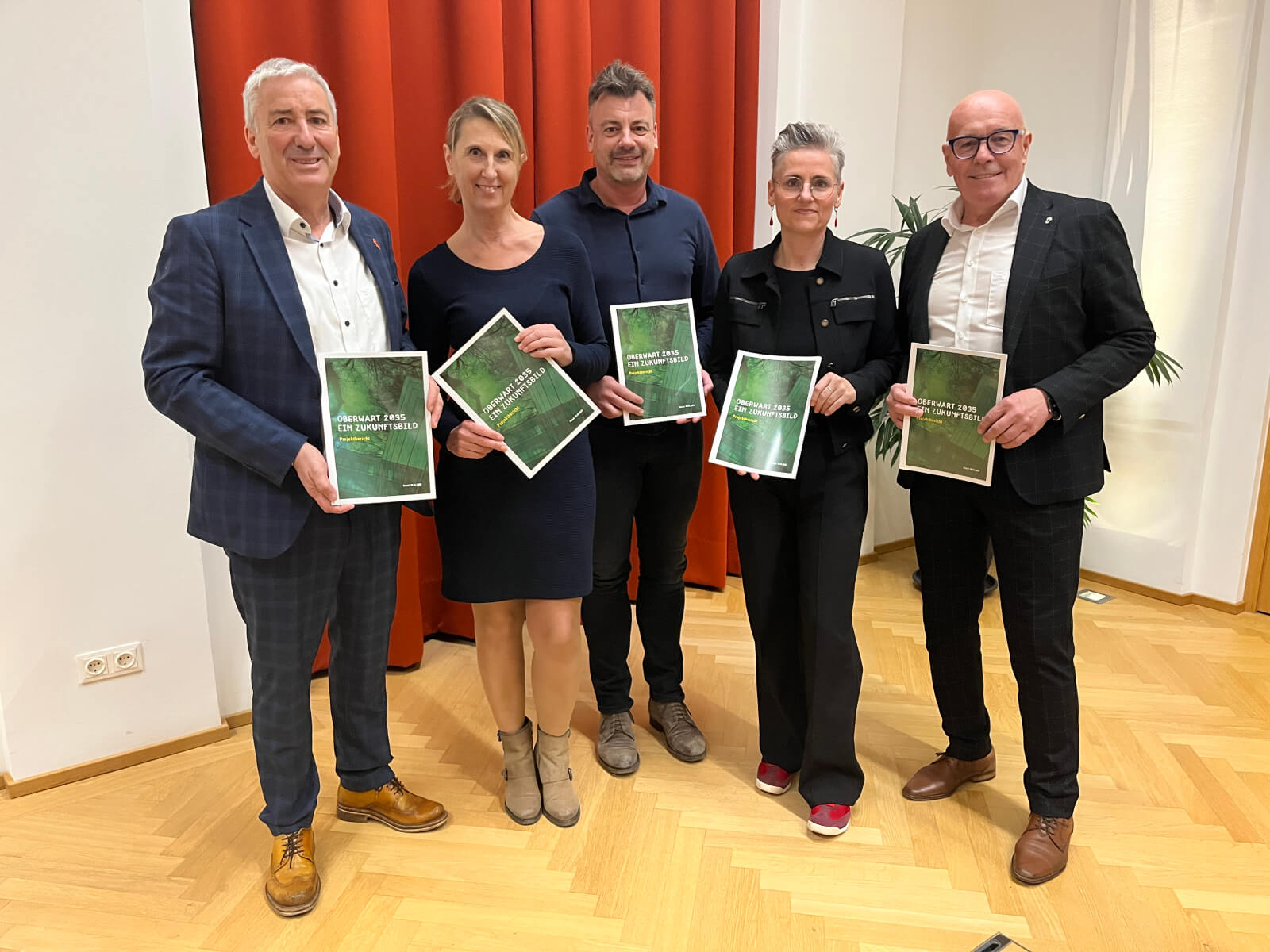 Eine Gruppe von fünf Personen hält Berichte mit dem Titel Oberwart 2035 – Ein Zukunftsbild vor rotem Vorhang.