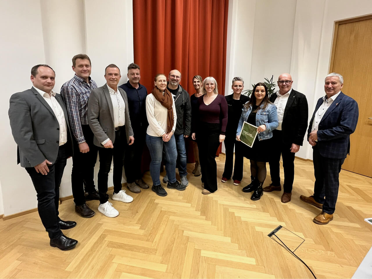 Gruppe von Menschen bei einem Event in einem Innenraum mit hölzernem Boden und roten Vorhängen, posierend und lächelnd.