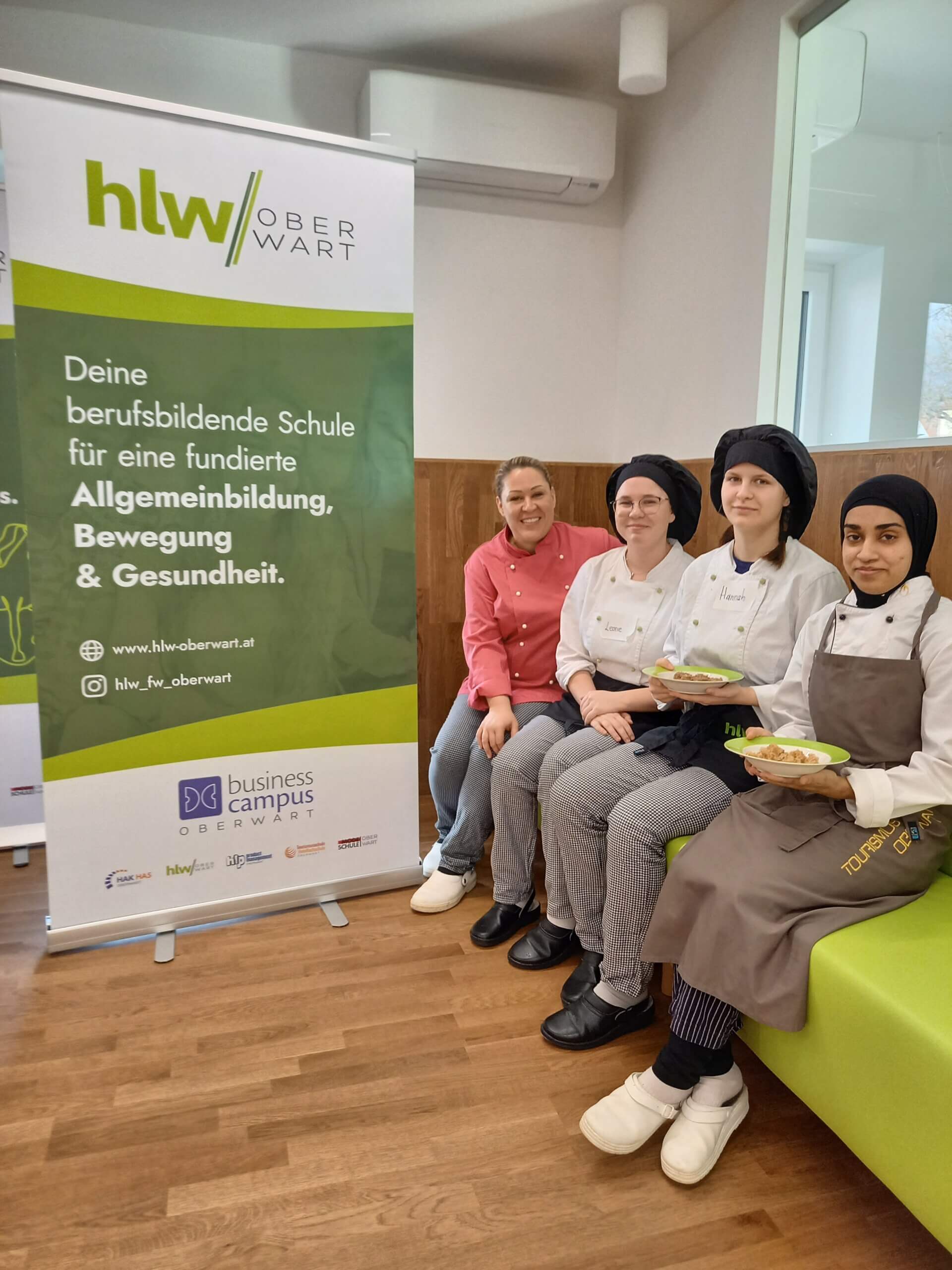 Schülerinnen der HLW Oberwart in Kochkleidung mit Tellern, neben einem Banner der Schule im Klassenzimmer.