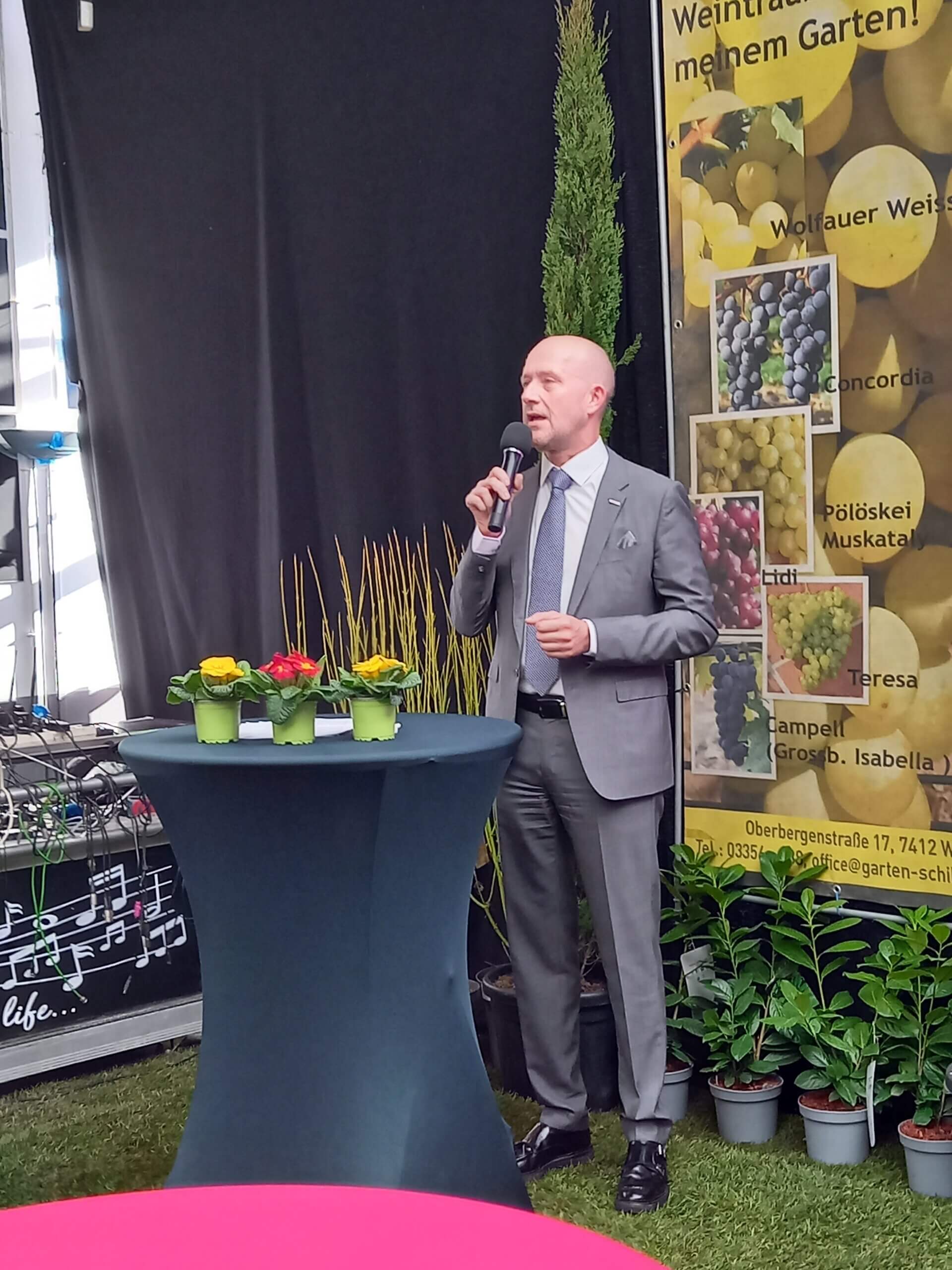 Mann im Anzug spricht bei einer Veranstaltung neben einem Tisch mit Blumen und einem Plakat über Traubensorten.