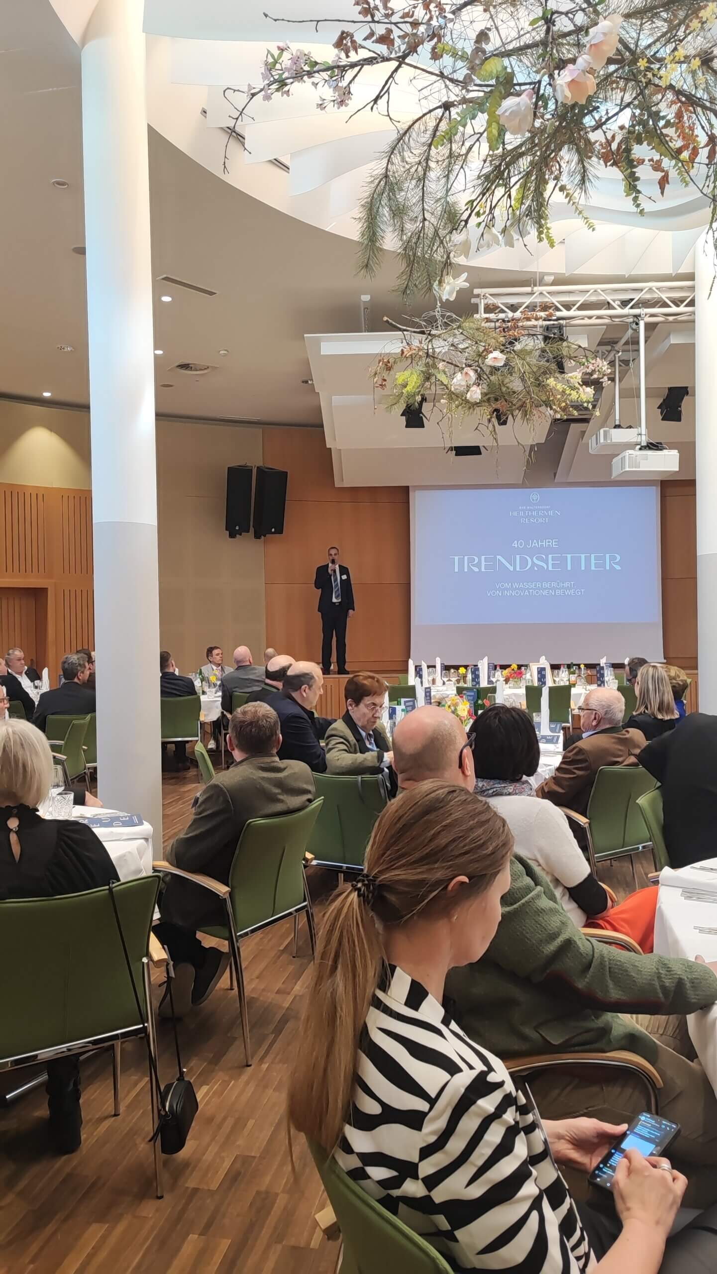 Konferenz im modernen Saal: Redner auf Bühne vor Publikum mit dekorativer Decke und Projektorbildschirm.