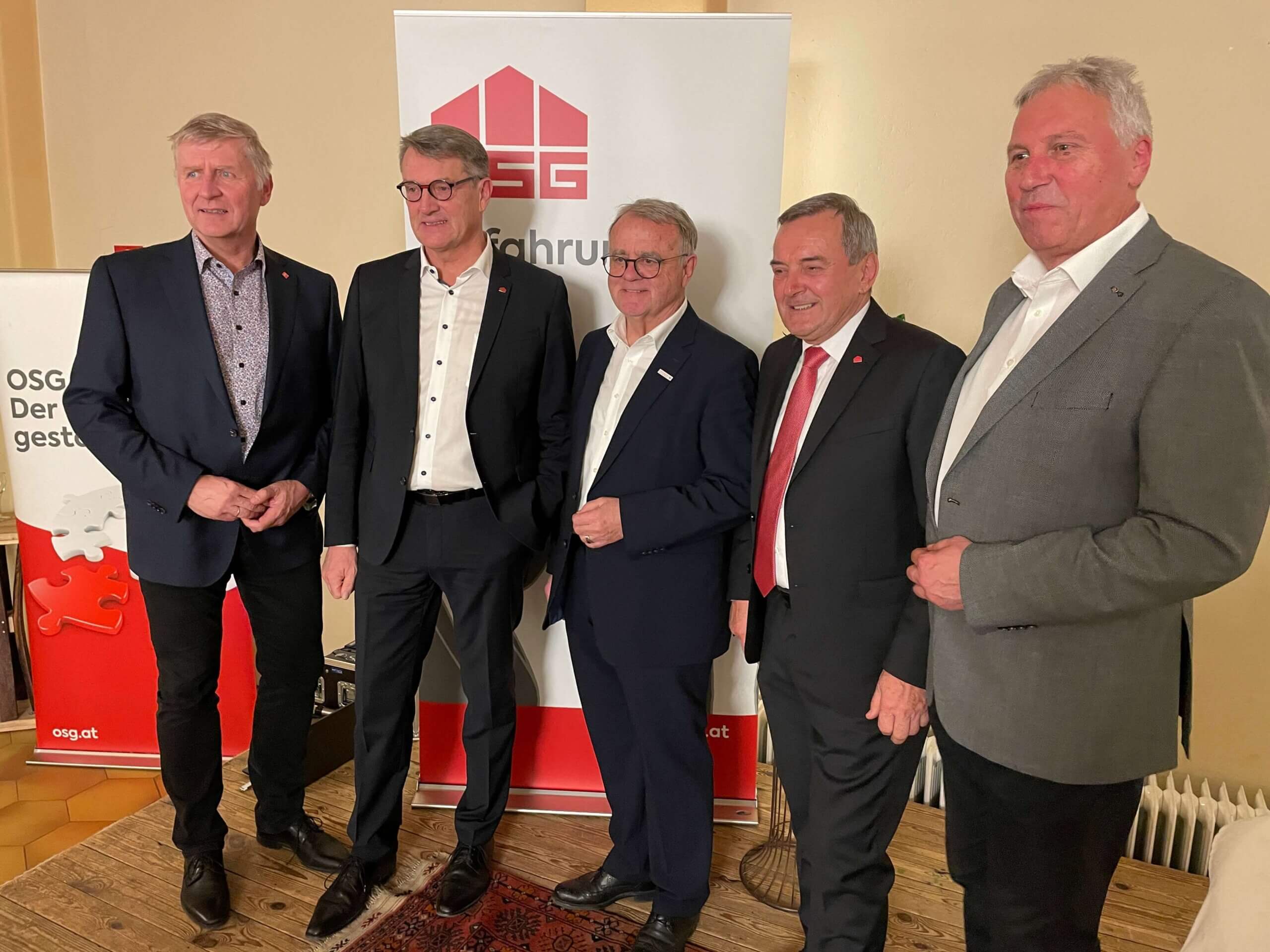 Männer in Anzügen posieren vor einem OSG-Banner bei einer Veranstaltung.