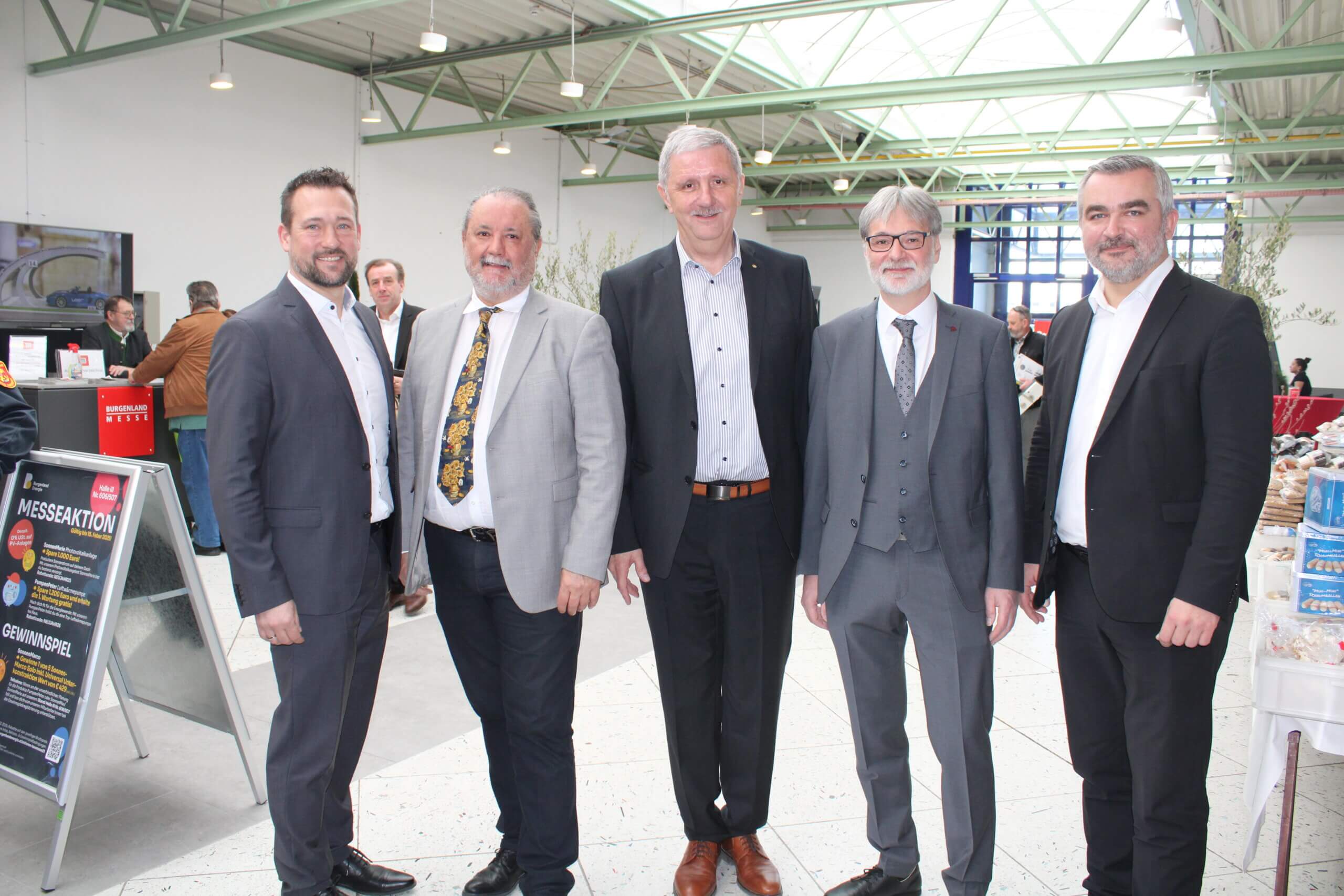 Gruppe von fünf Männern in Anzügen auf einer Messe, vor einem Infostand mit Plakaten und Produkten im Hintergrund.