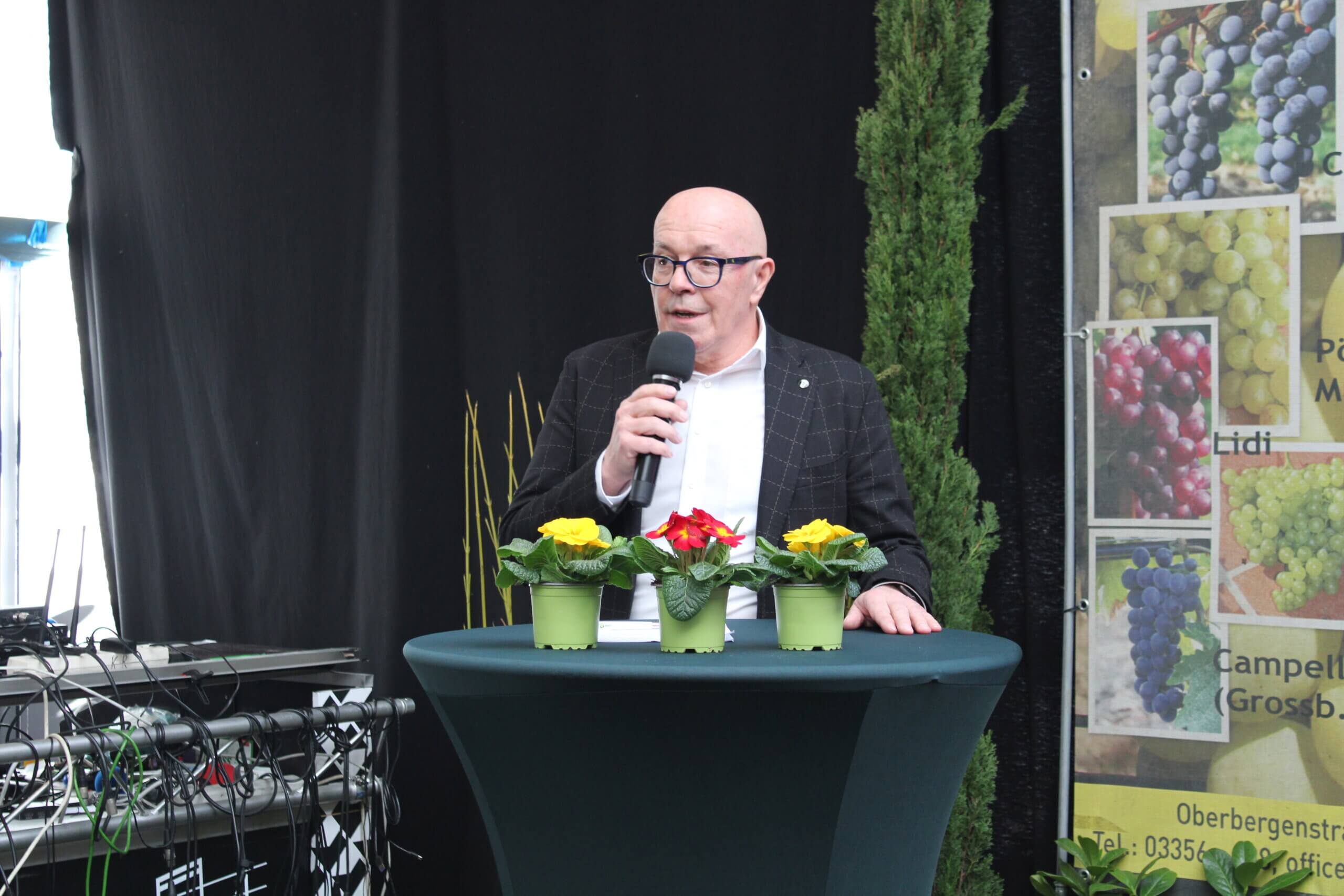 Ein Redner mit Mikrofon auf einer Bühne, Tisch mit Blumen, Hintergrund mit Weinrebenposter.