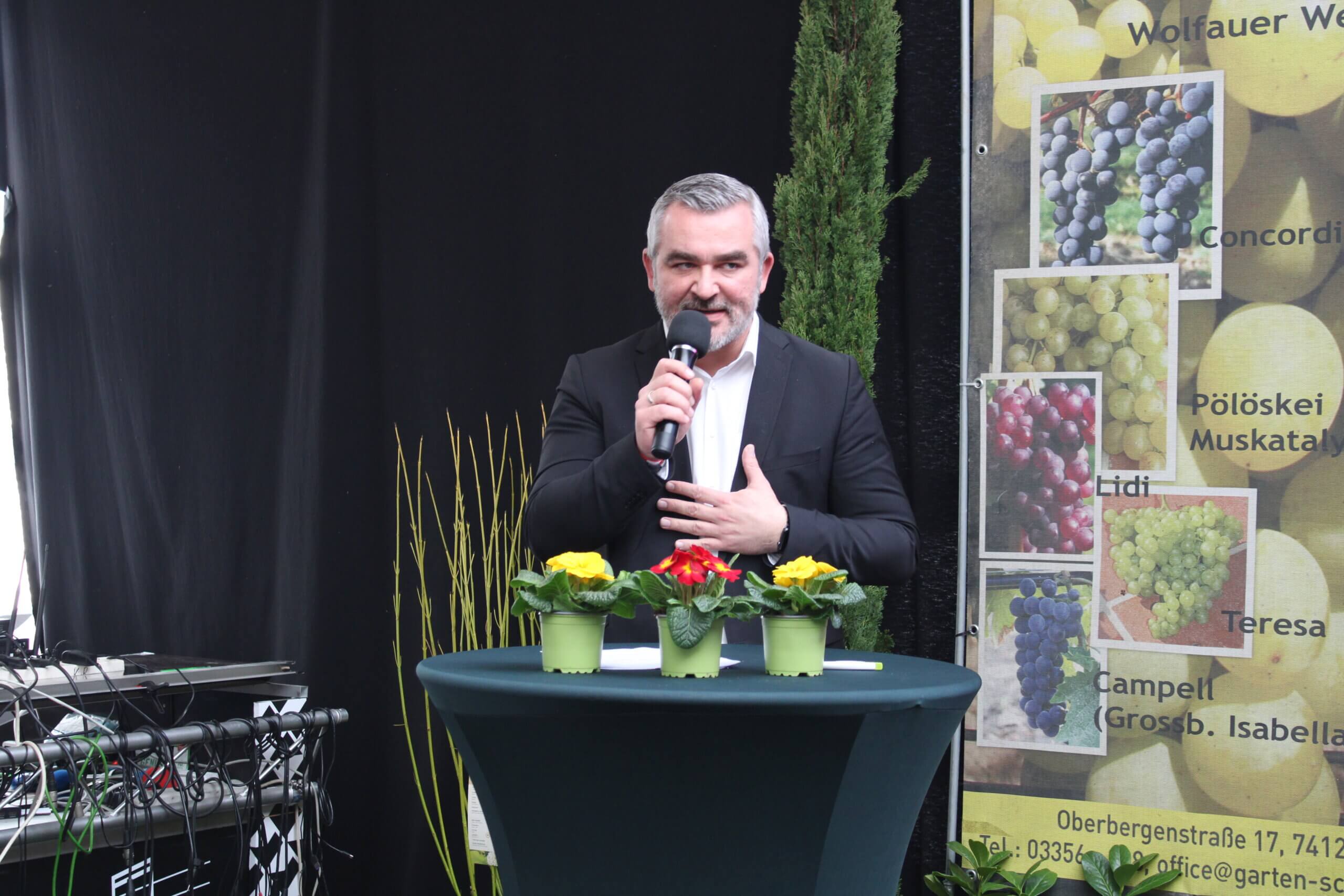Mann hält Rede auf Gartenveranstaltung mit Blumen und Weinreben-Poster im Hintergrund.