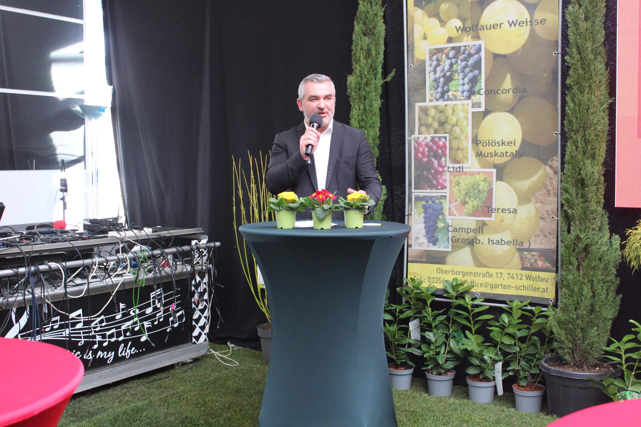 Ein Mann hält eine Rede auf einer Veranstaltung, Dekoration mit Pflanzen und Poster über Rebsorten im Hintergrund.