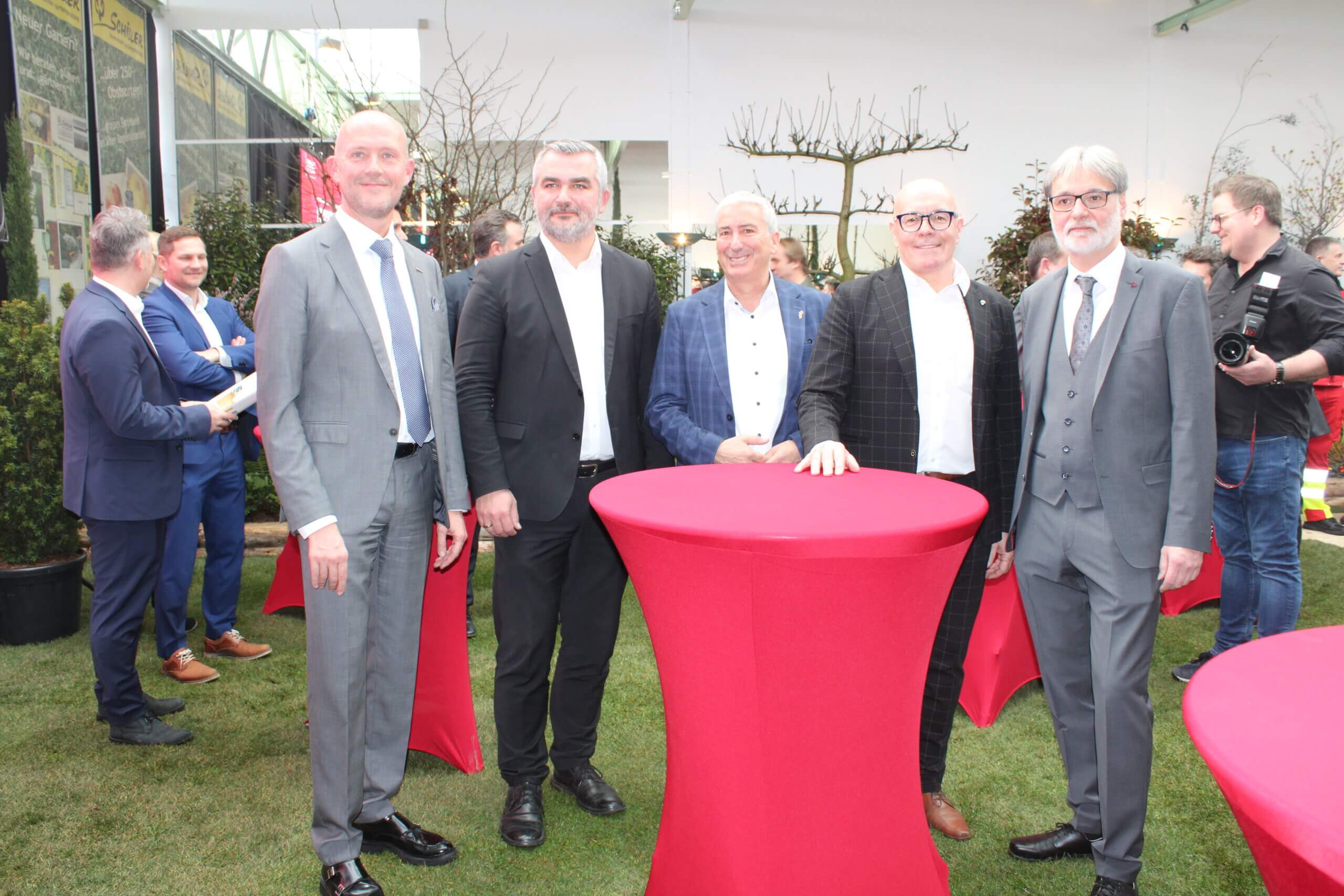 Fünf Männer in Anzügen bei einer Veranstaltung im Freien, stehend an runden Tischen mit roten Tischdecken.