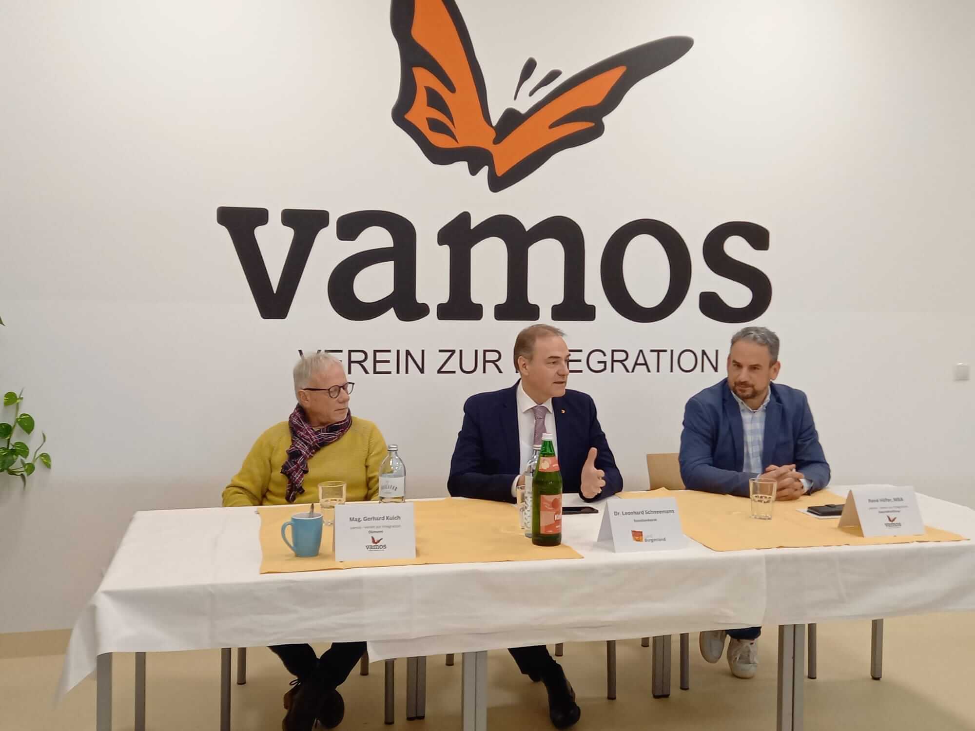 Drei Männer bei einer Diskussionsveranstaltung von Vamos Verein zur Integration an einem Tisch mit Logo im Hintergrund.