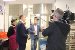 Führung durch Industrieküche: Chef erklärt Equipment, Kamerateam filmt, Mitarbeiter und Besucher im Gespräch.