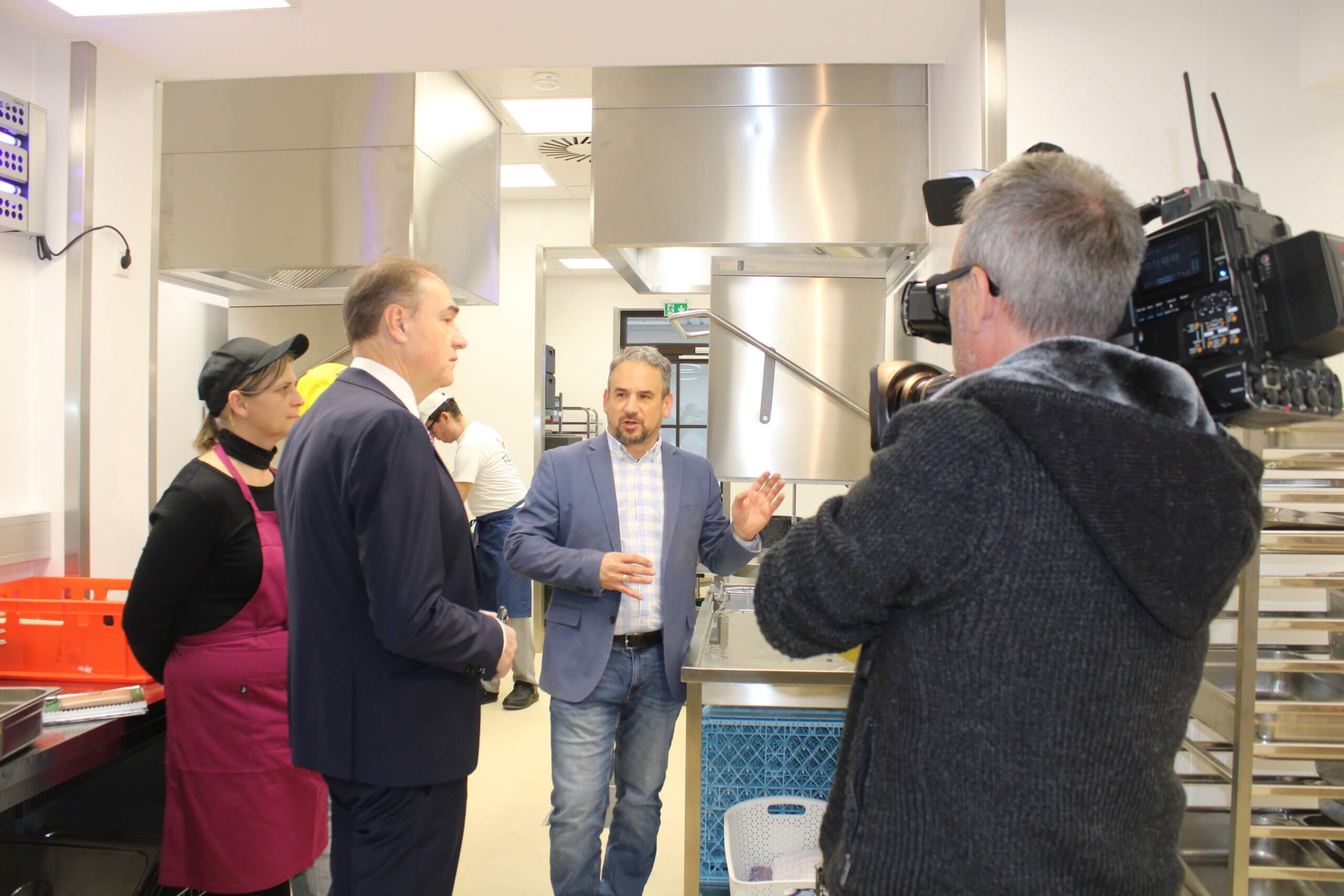 Führung durch Industrieküche: Chef erklärt Equipment, Kamerateam filmt, Mitarbeiter und Besucher im Gespräch.