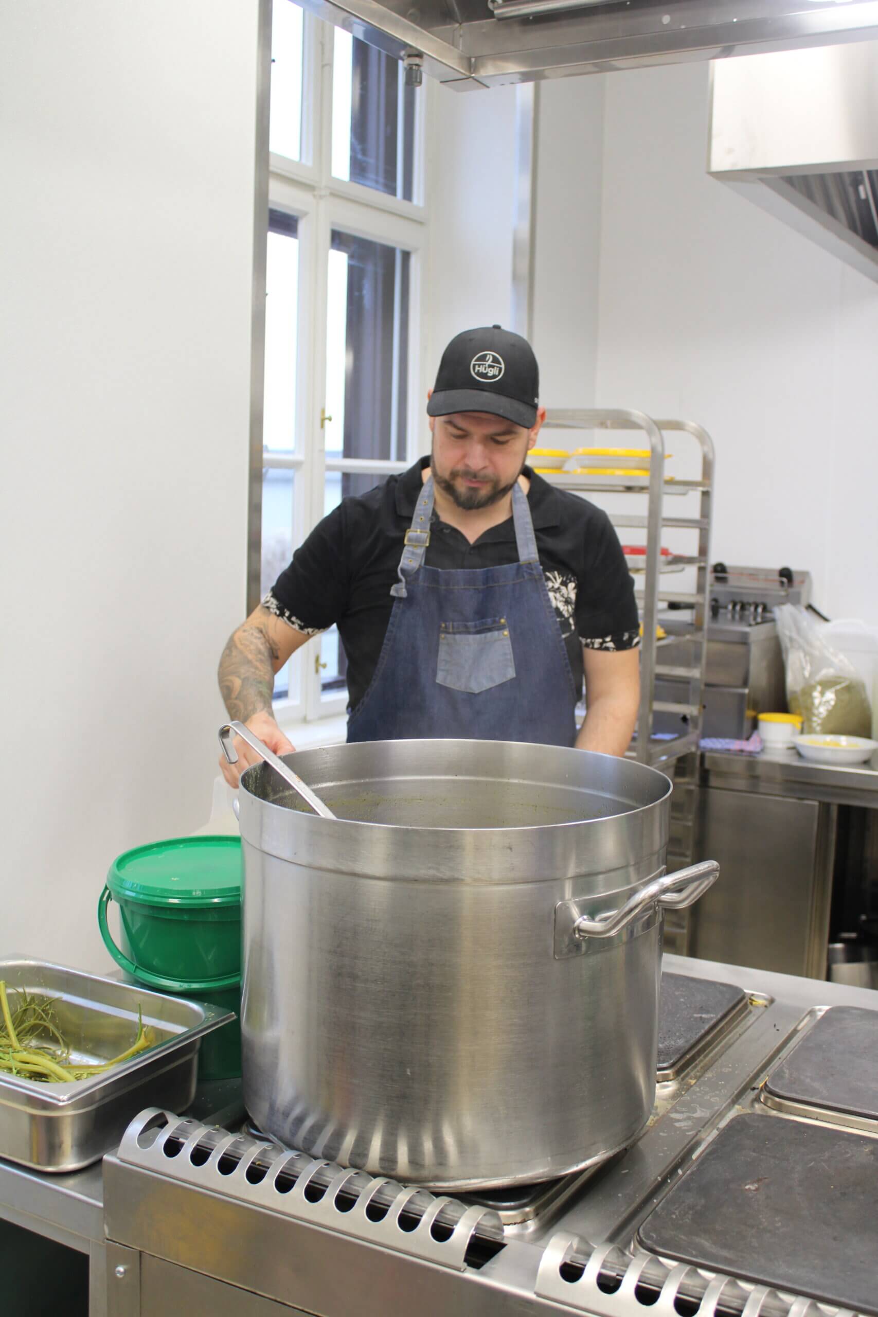 Koch in schwarzem T-Shirt und Schürze rührt in großem Topf in moderner Küche.