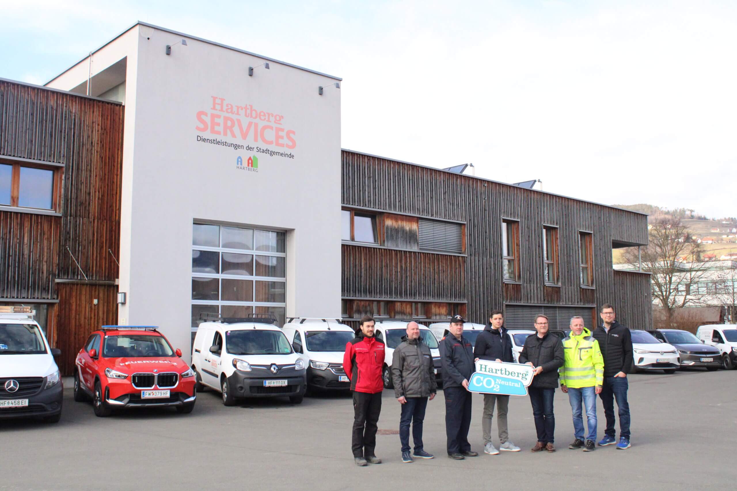 Gruppe vor Hartberg Services Gebäude mit CO2-neutralem Schild, umgeben von Firmenfahrzeugen.