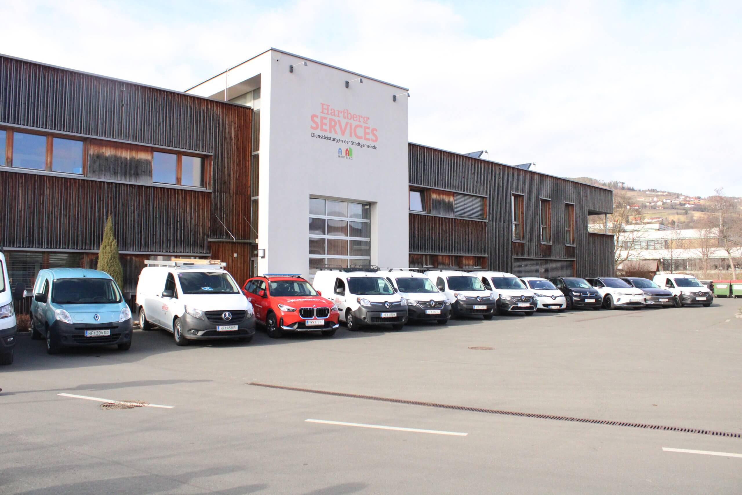 Mehrere Fahrzeuge parken vor dem modernen Gebäude von Hartberg Services an einem sonnigen Tag.