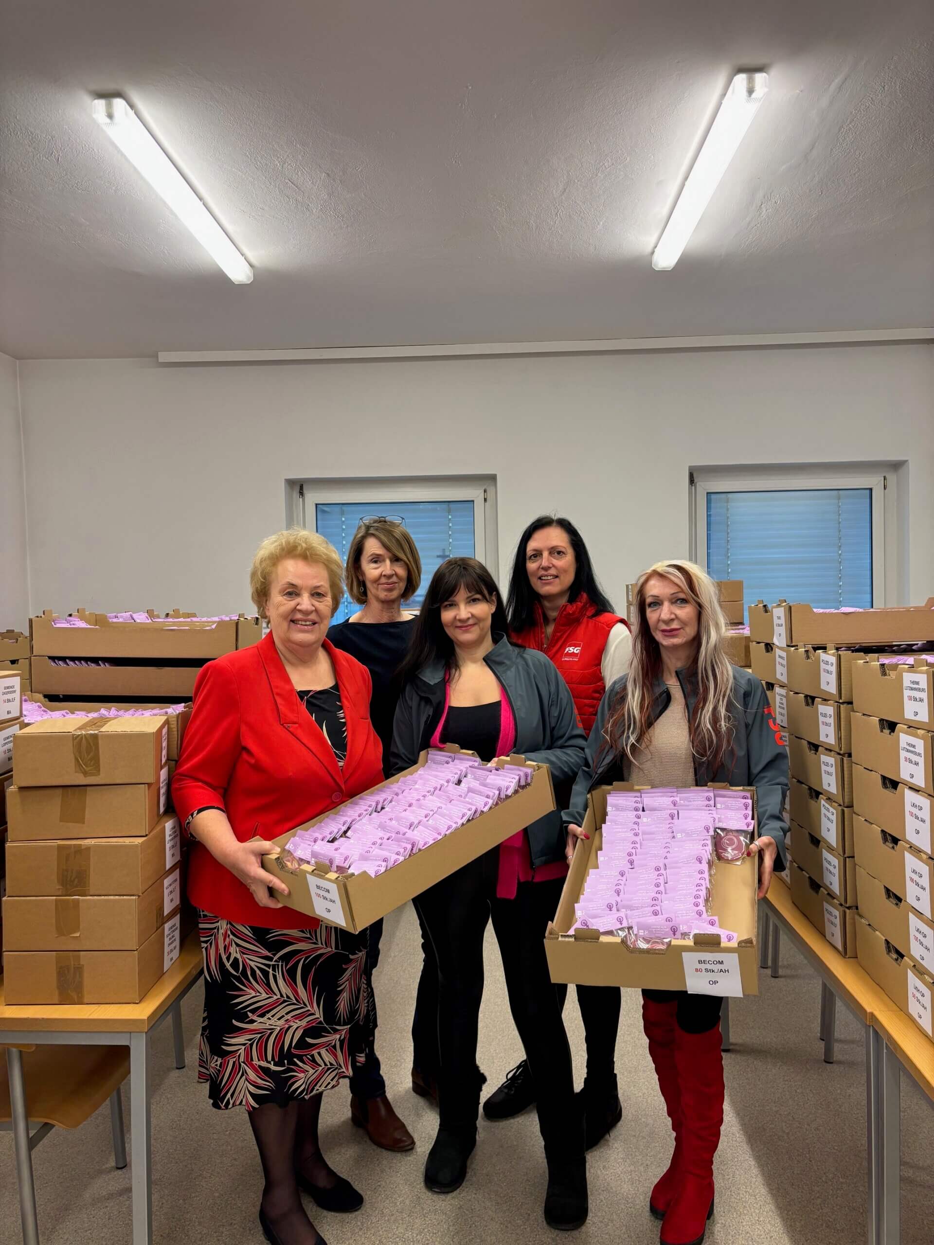 Gruppe von Frauen mit Kartons voller Produkte, lächelnd in Lagerhalle, Zusammenarbeit und Teamgeist symbolisierend.