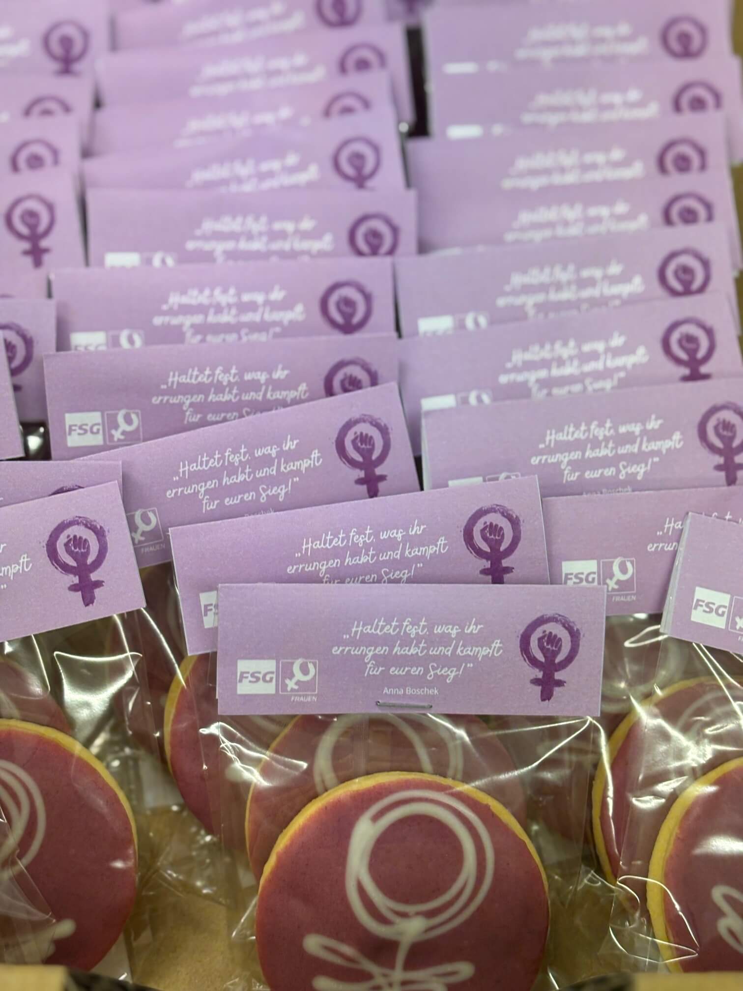 Lila Kekse mit feministischen Symbolen und motivierender Botschaft für Frauenrechte, in durchsichtigen Verpackungen präsentiert.