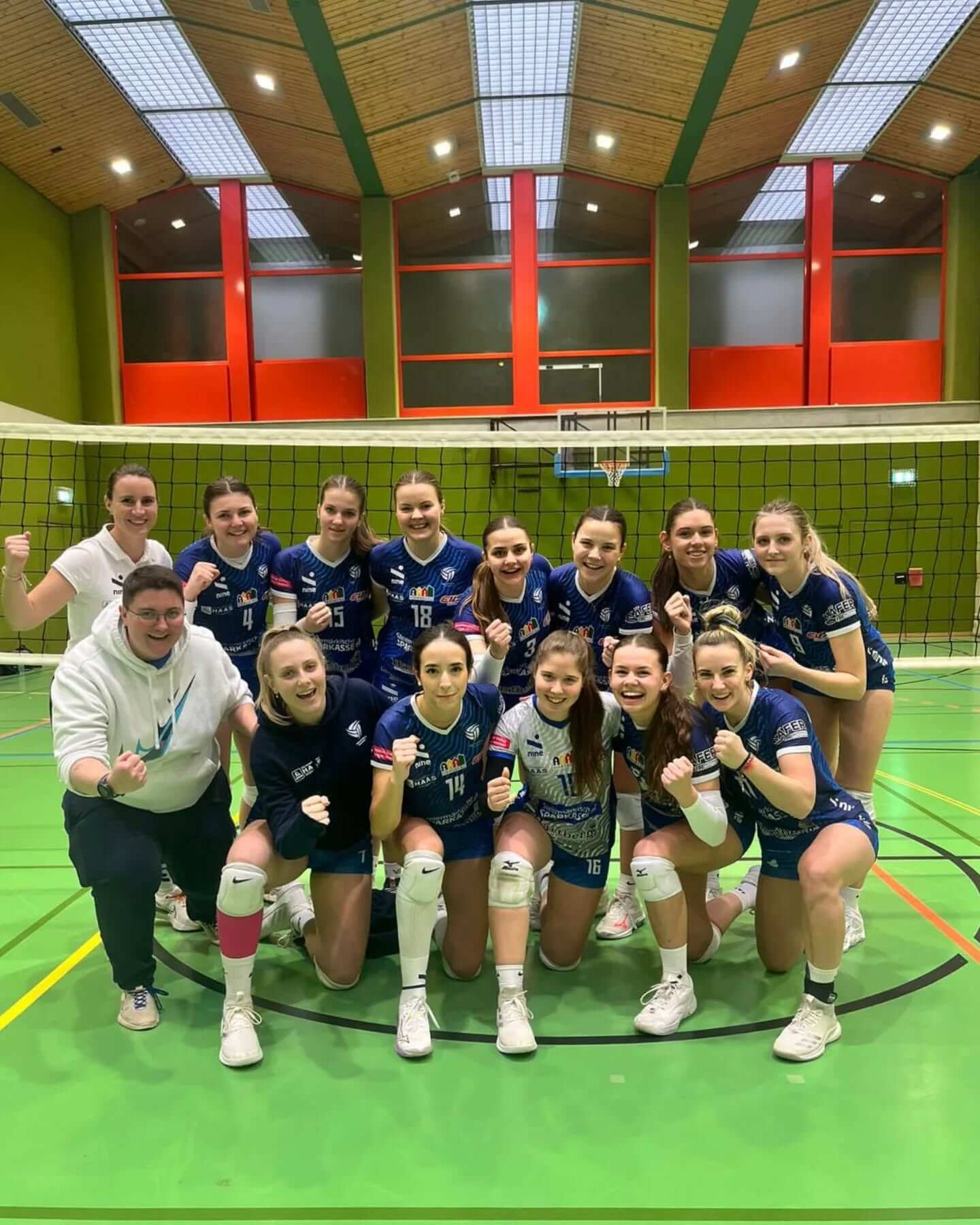 Frauen-Volleyballteam jubelt in Sporthalle vor dem Netz, zeigt Teamgeist und Freude am Spielen.