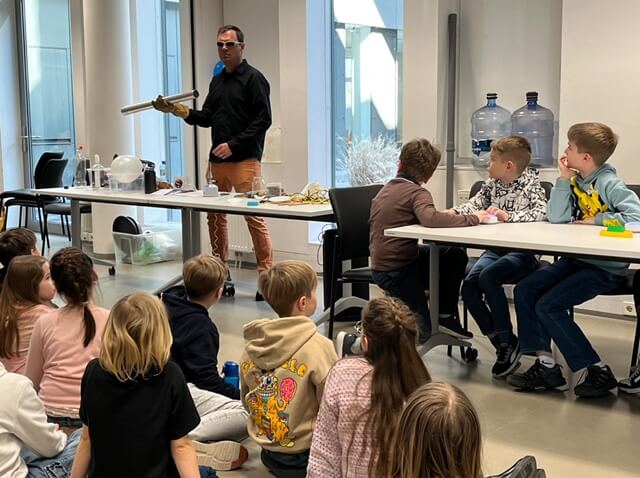 Mann führt wissenschaftliches Experiment vor Kindern durch, die aufmerksam in einem Schulraum sitzen.