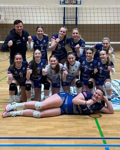 Frauen-Volleyball-Team feiert mit ihrem Trainer im Sportzentrum vor dem Netz. Teamgeist und Freude im Vordergrund.
