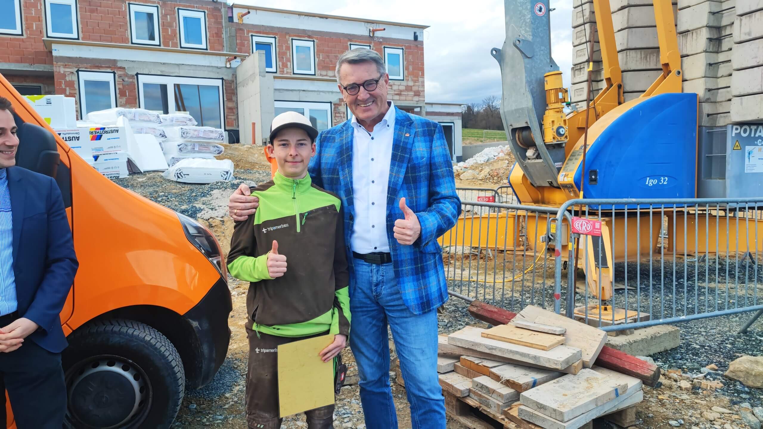 Personen posieren auf einer Baustelle neben einem Baukran, mit Daumen hoch und einem orangefarbenen Fahrzeug im Hintergrund.