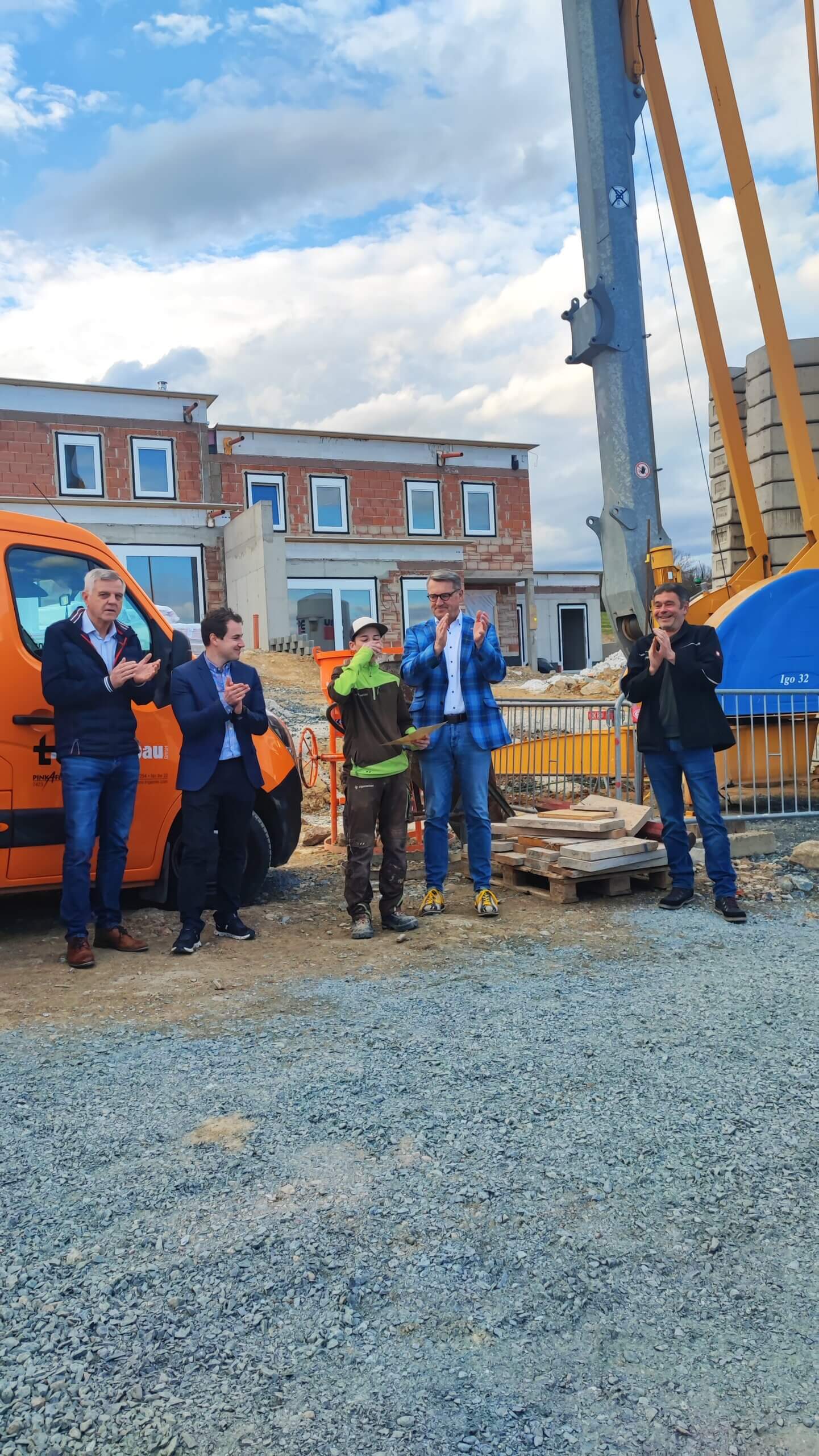 Gruppe applaudiert bei einer Baustellenveranstaltung vor einem orangefarbenen Transporter und einem Rohbau.