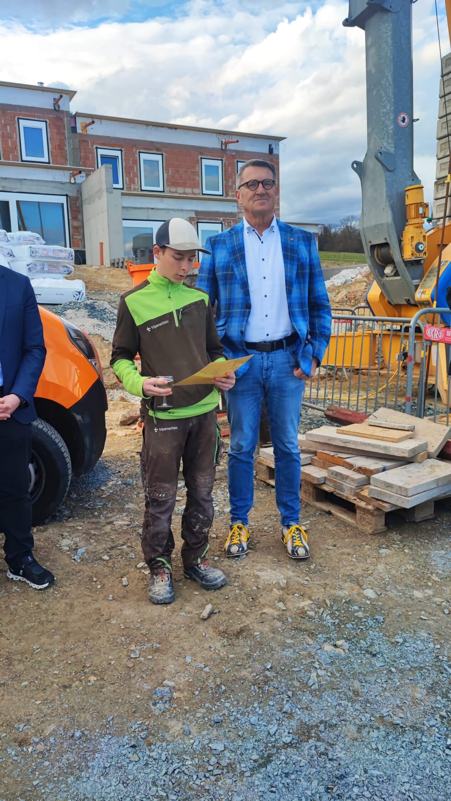 Zwei Personen auf einer Baustelle, ein Mann im Anzug und ein Jugendlicher in Arbeitskleidung mit einem Blatt Papier.