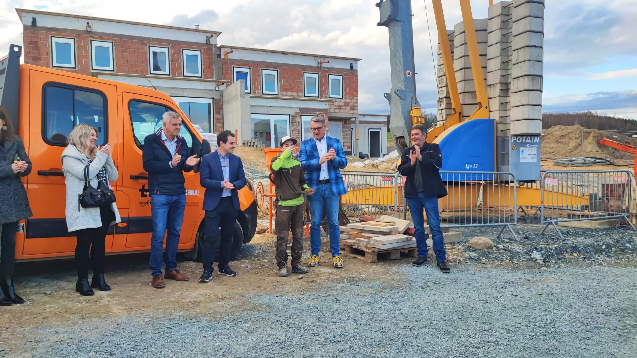 Gruppe von Personen applaudiert bei einer Baustelle mit orangem Fahrzeug und Kran im Hintergrund.