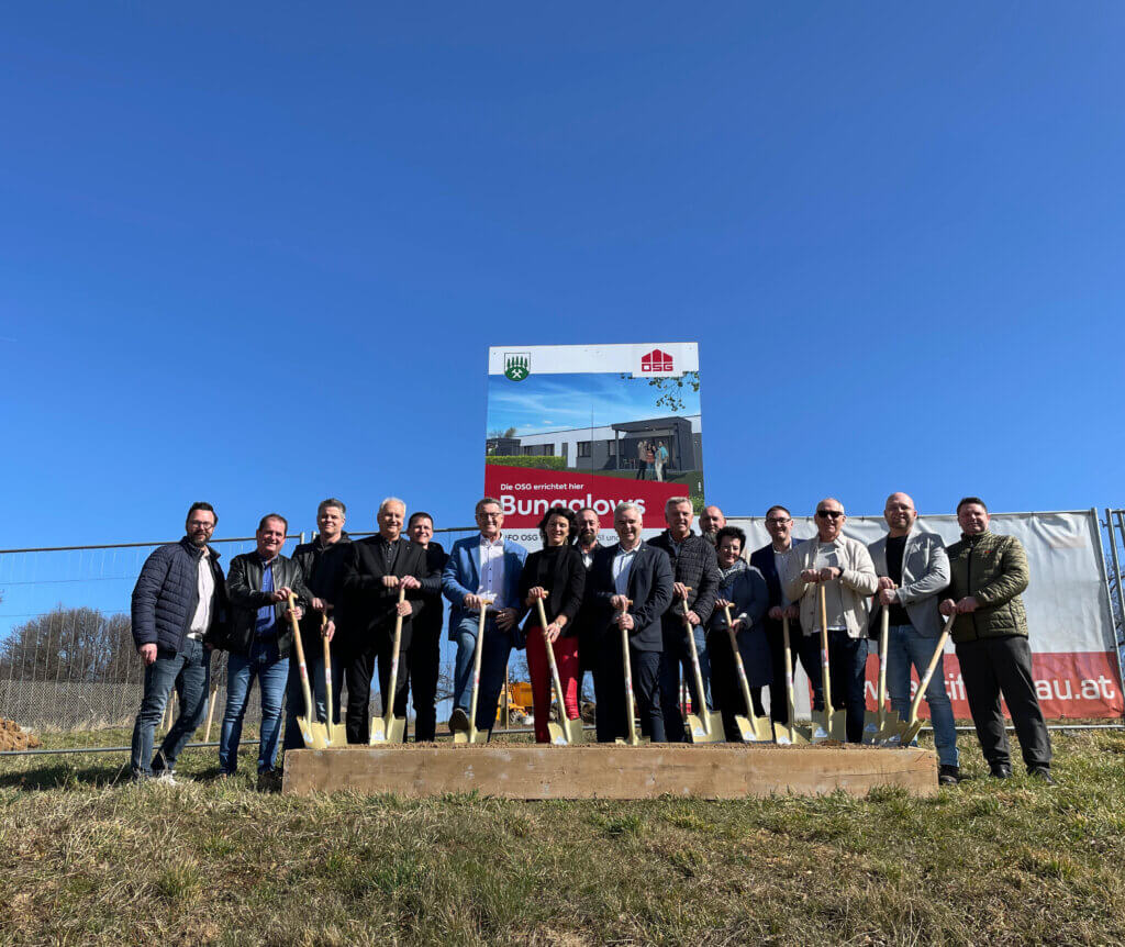 Gruppe von Personen beim Spatenstich für ein neues Bauprojekt vor einem Werbeschild unter blauem Himmel.