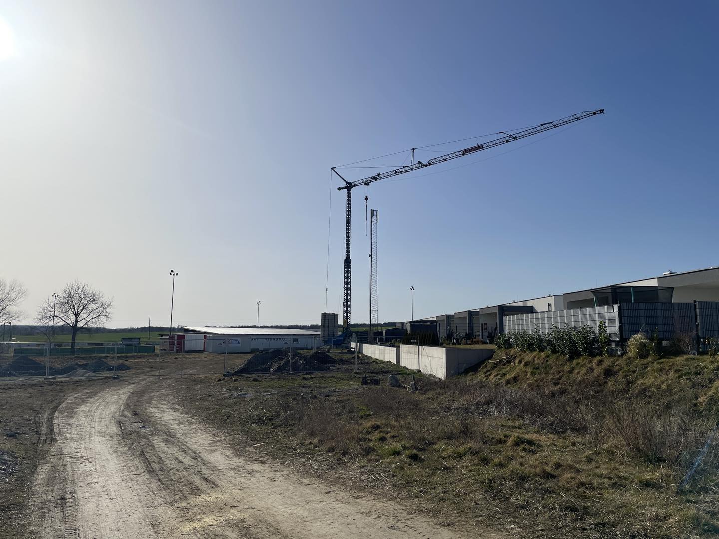 Kran auf Baustelle mit blauem Himmel und Gebäuden im Hintergrund, sonniger Tag, Weg im Vordergrund.