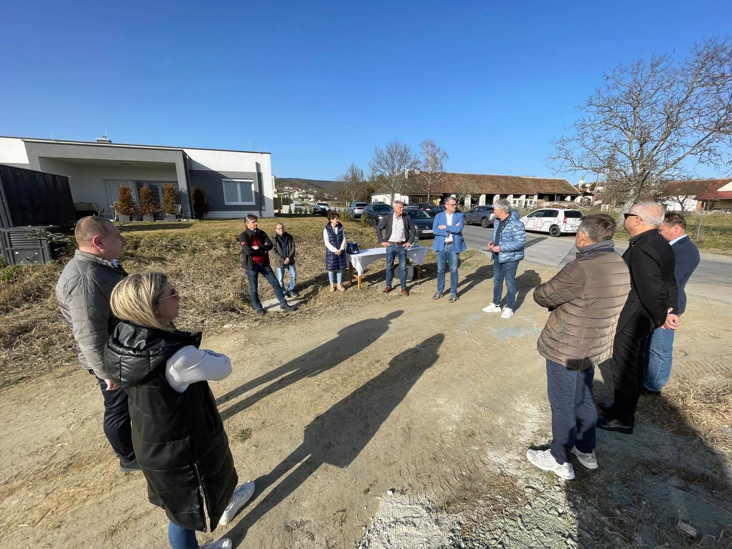 Gruppe von Menschen trifft sich bei sonnigem Wetter im Freien vor modernen Gebäuden und Autos.