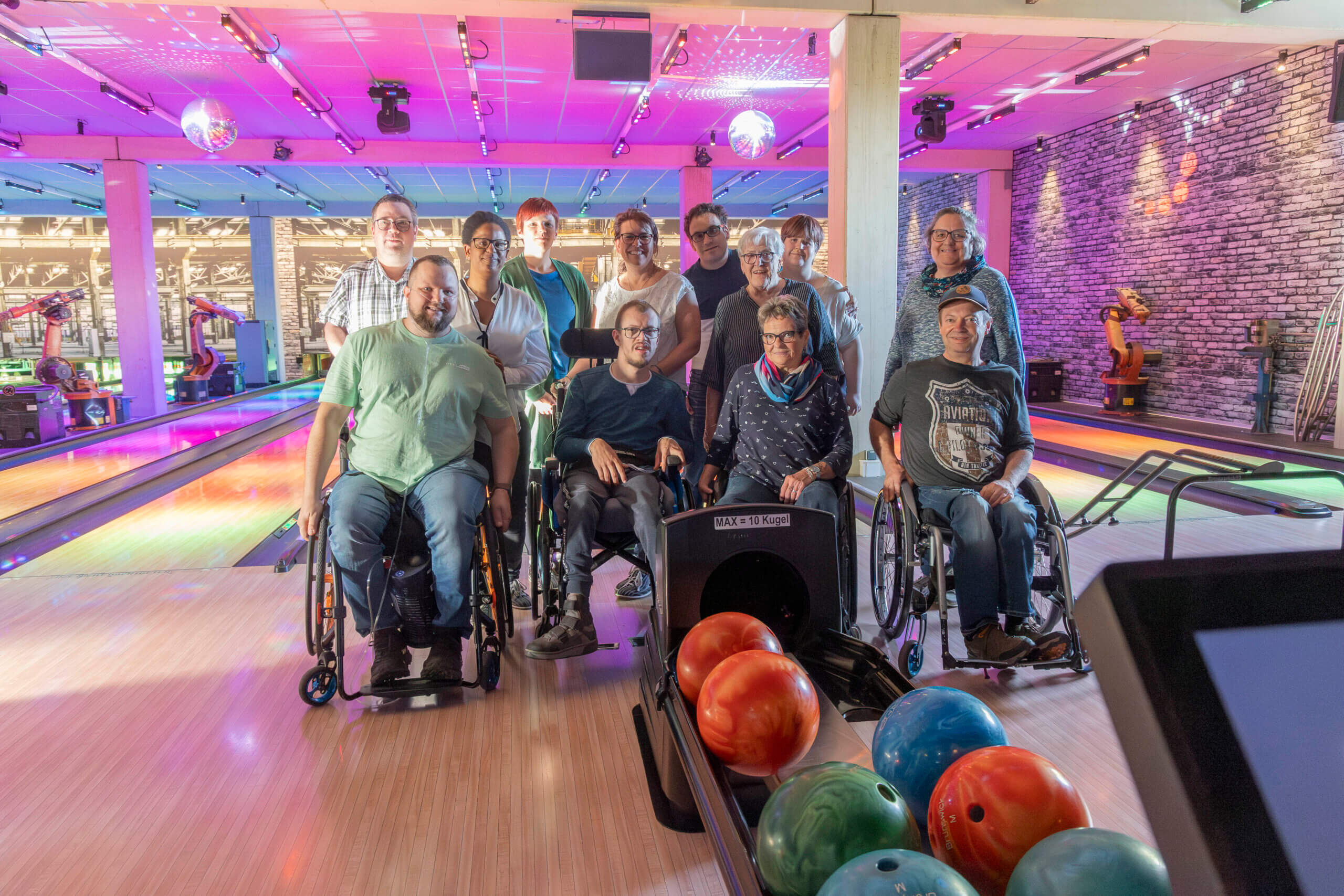 Gruppe von Menschen im Rollstuhl beim Bowling in einer bunt beleuchteten Halle, lächelnd für ein Gruppenfoto.