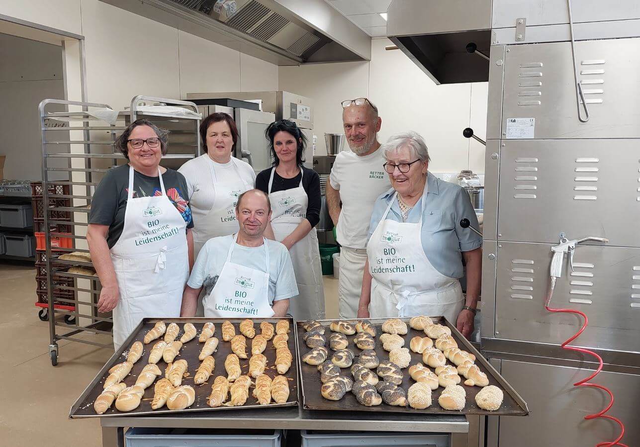 Bäcker-Team präsentiert frische Bio-Brötchen in Backstube, alle tragen Schürzen mit Aufschrift BIO ist meine Leidenschaft.