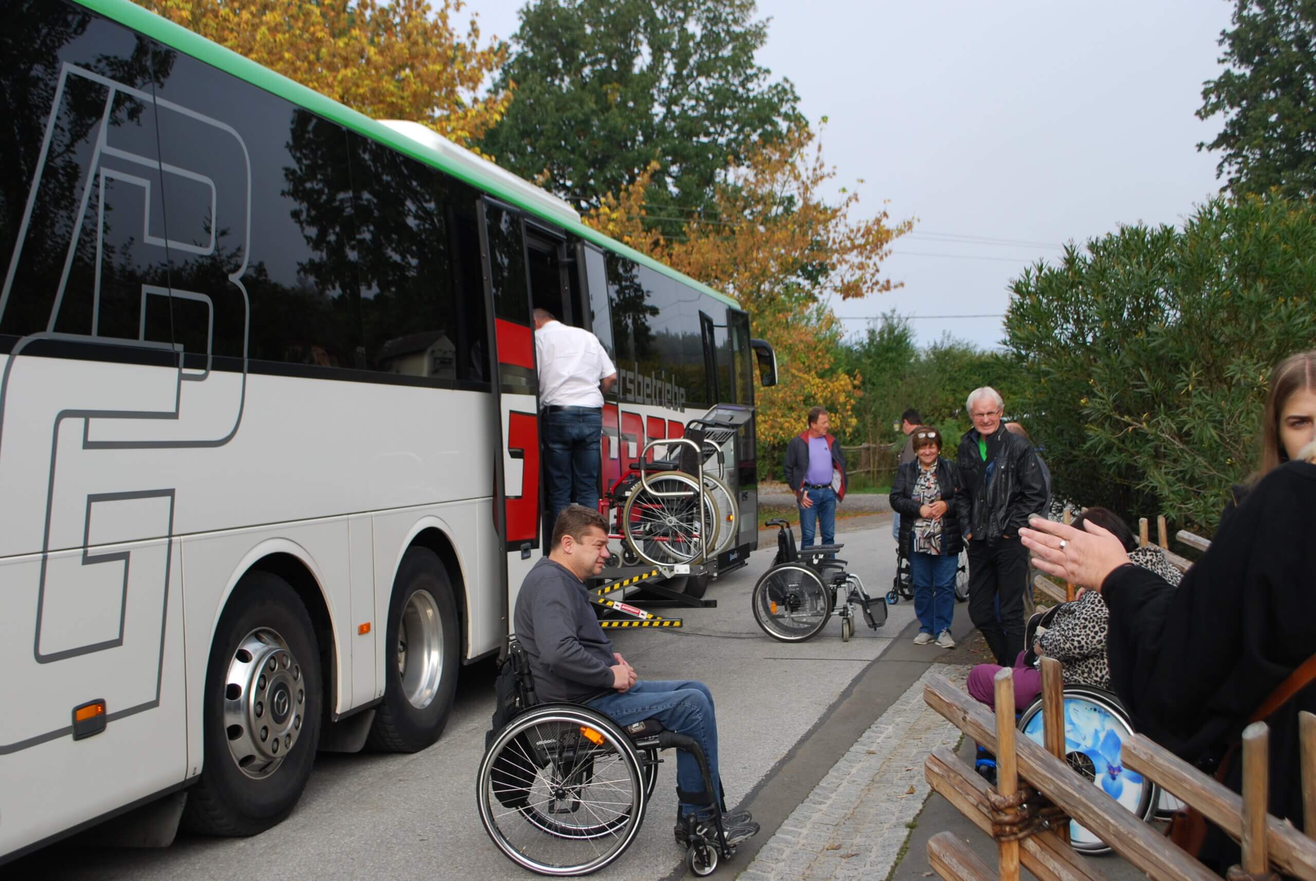 Menschen steigen in Bus ein, Rollstuhlfahrer warten auf Einstiegshilfe, barrierefreies Reisen im Herbst.