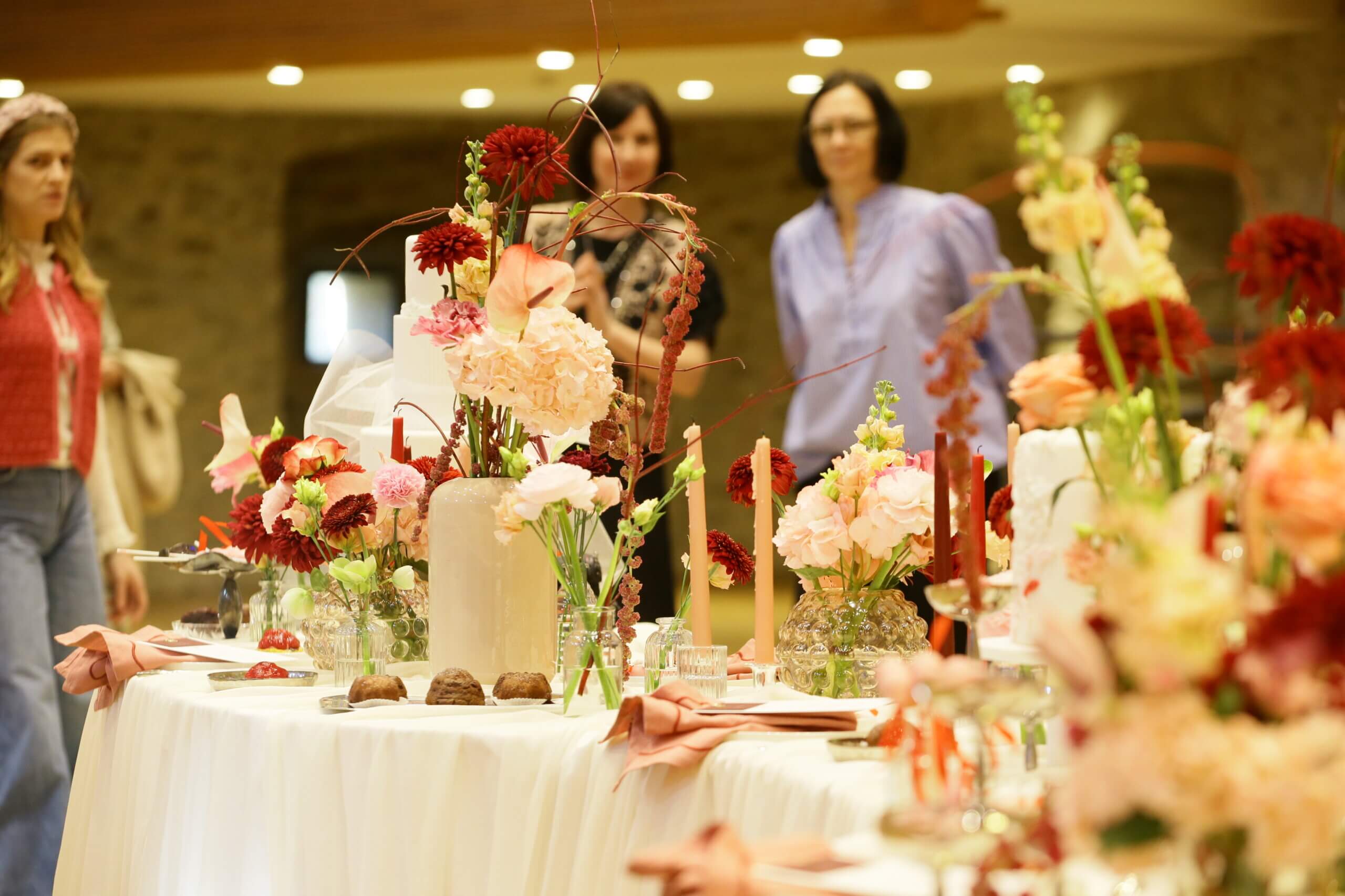 Elegant gedeckter Tisch mit üppigen Blumengestecken, Kerzen und Dekorationen, betrachtet von drei Personen im Hintergrund.