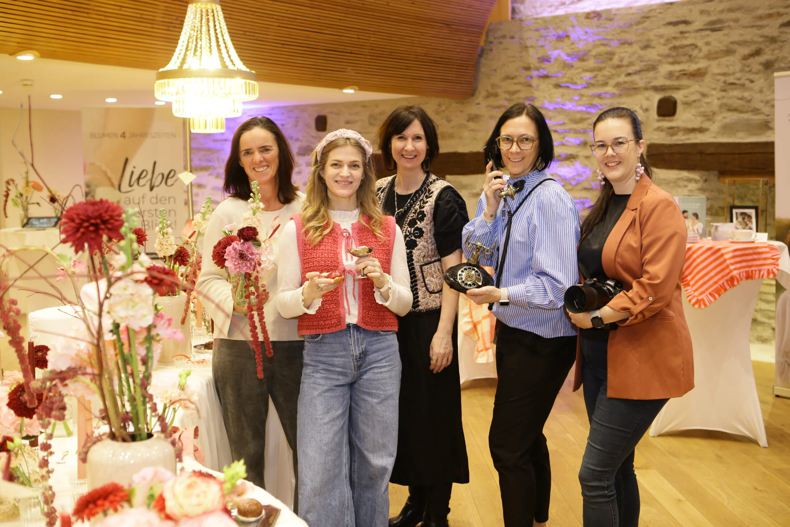 Fünf Frauen lächeln in die Kamera, umgeben von Blumenarrangements und Dekorationen in einem festlichen Raum.