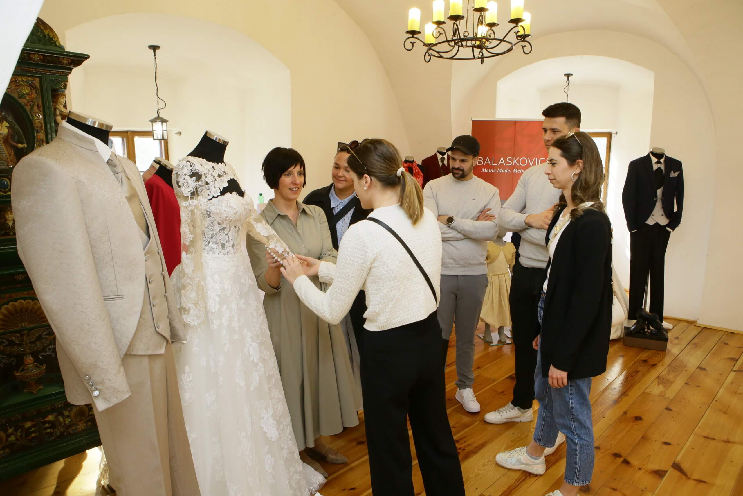 Gruppe betrachtet Brautmode und Anzüge bei Modenschau-Event in elegantem Ausstellungsraum.