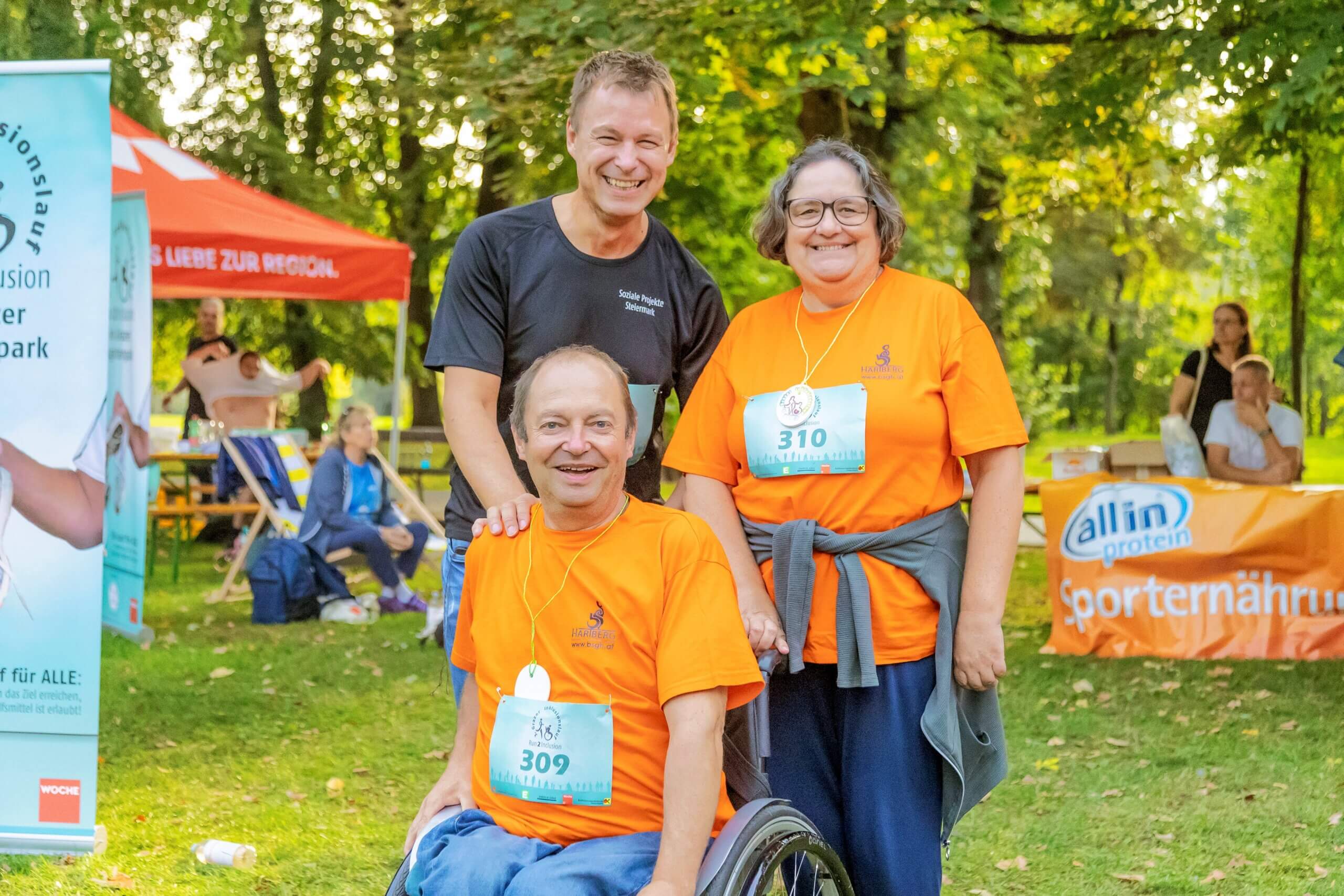 Drei Menschen in Orange bei einem inklusiven Lauf-Event im Park, lächelnd und in sportlicher Atmosphäre.
