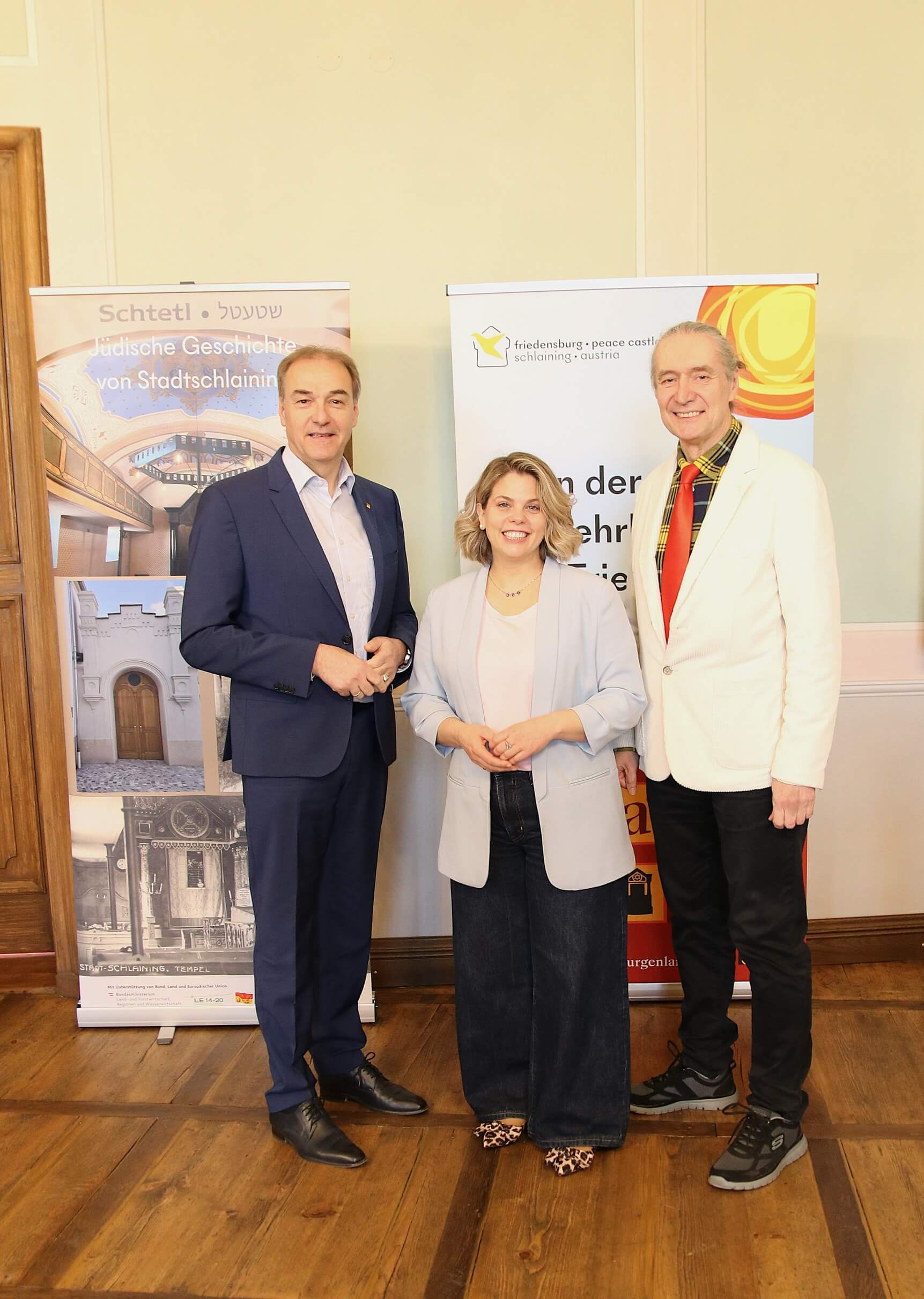 Personen vor Bannern im Friedenszentrum Schlaining, Österreich, lächeln bei Veranstaltung. Holzboden und historische Kulisse.