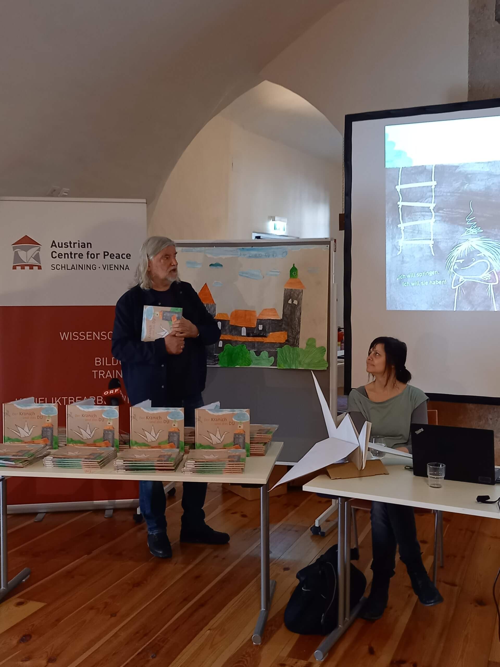 Autor präsentiert Buch im Austrian Centre for Peace. Frau beobachtet. Bücherstapel und Origami-Kranich auf Tischen.