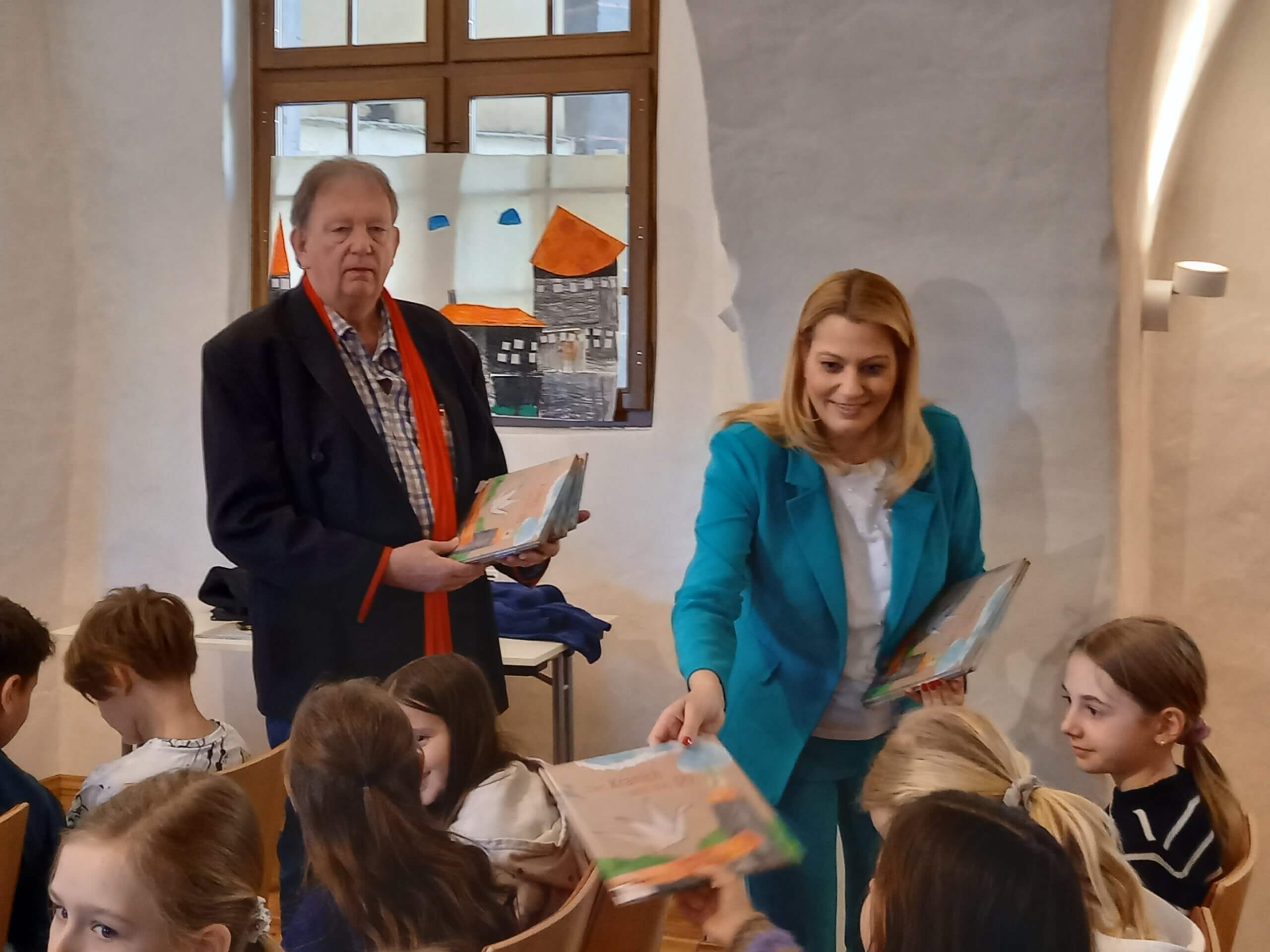 Zwei Erwachsene verteilen Bücher an Kinder in einem hellen Raum mit Kunstwerken an den Fenstern.