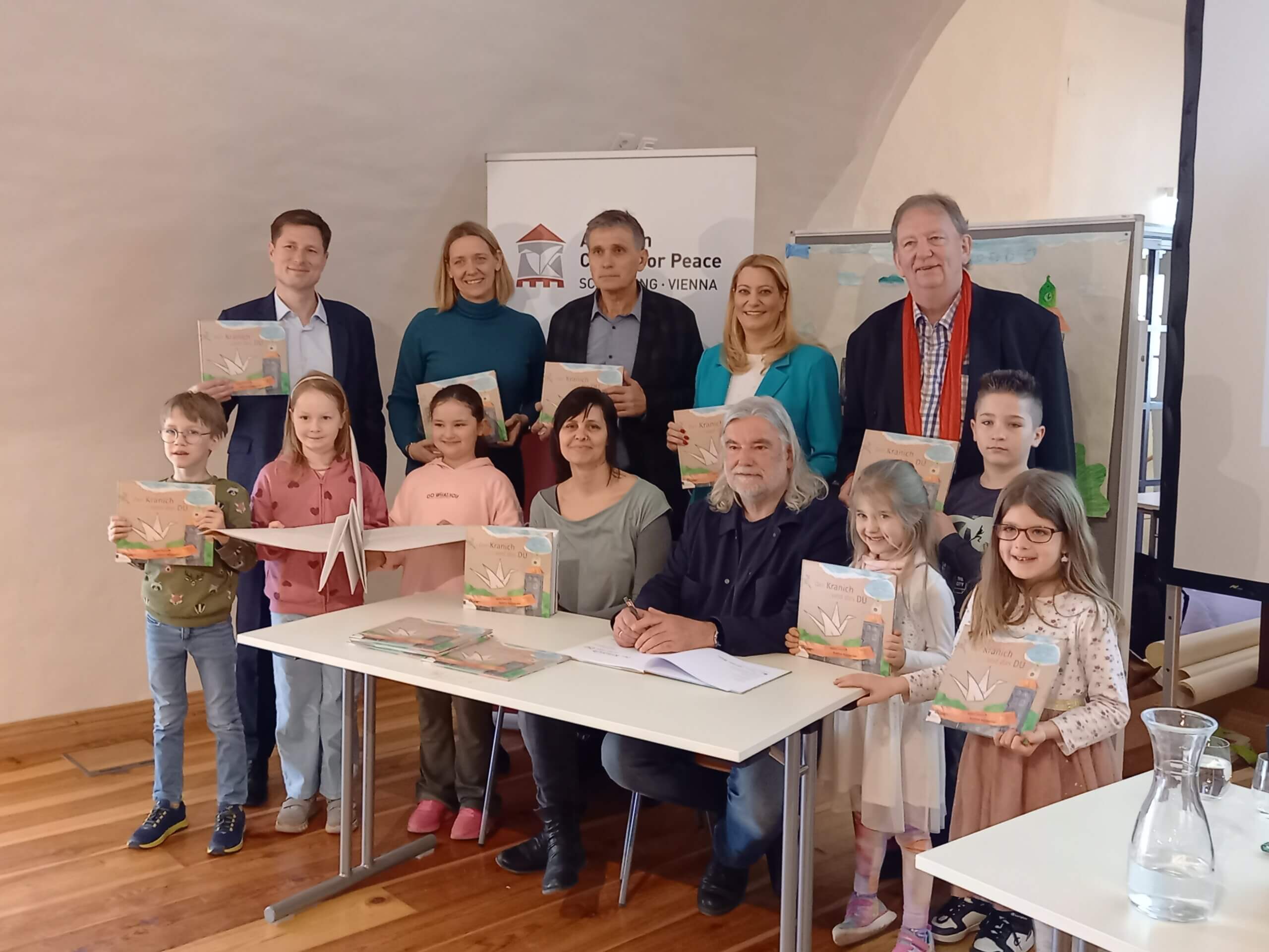 Gruppe bei Buchpräsentation: Erwachsene und Kinder mit Büchern, lächelnd vor einem Banner, in einem Veranstaltungsraum.