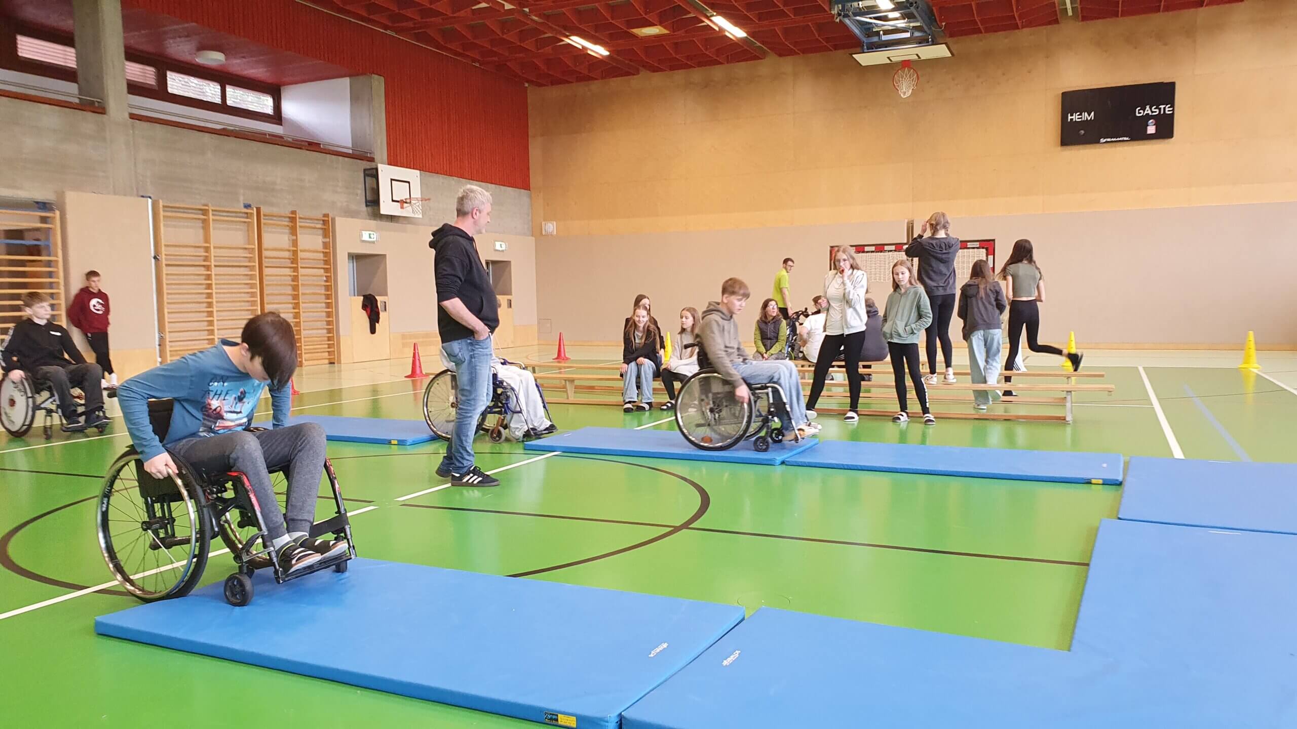 Menschen im Rollstuhl bei Sportaktivität in Turnhalle, mit Hindernisparcours und Zuschauern. Barrierefreiheit erleben.