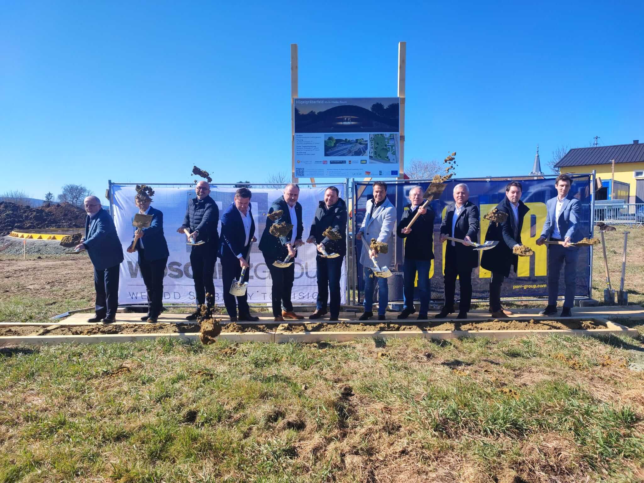 Personengruppe beim Spatenstich für ein Bauprojekt, sonniger Tag, Banner im Hintergrund.