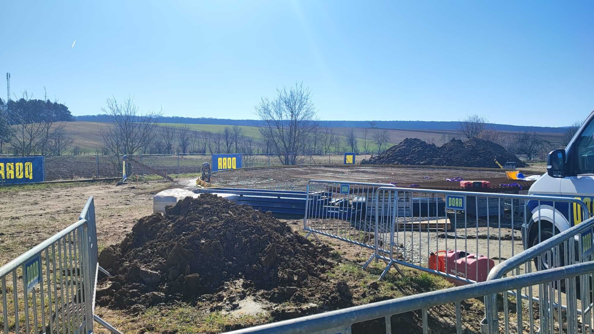 Baustelle auf freiem Feld mit Erdarbeiten, Bauzäunen und Fahrzeug bei sonnigem Wetter, ländliche Landschaft im Hintergrund.