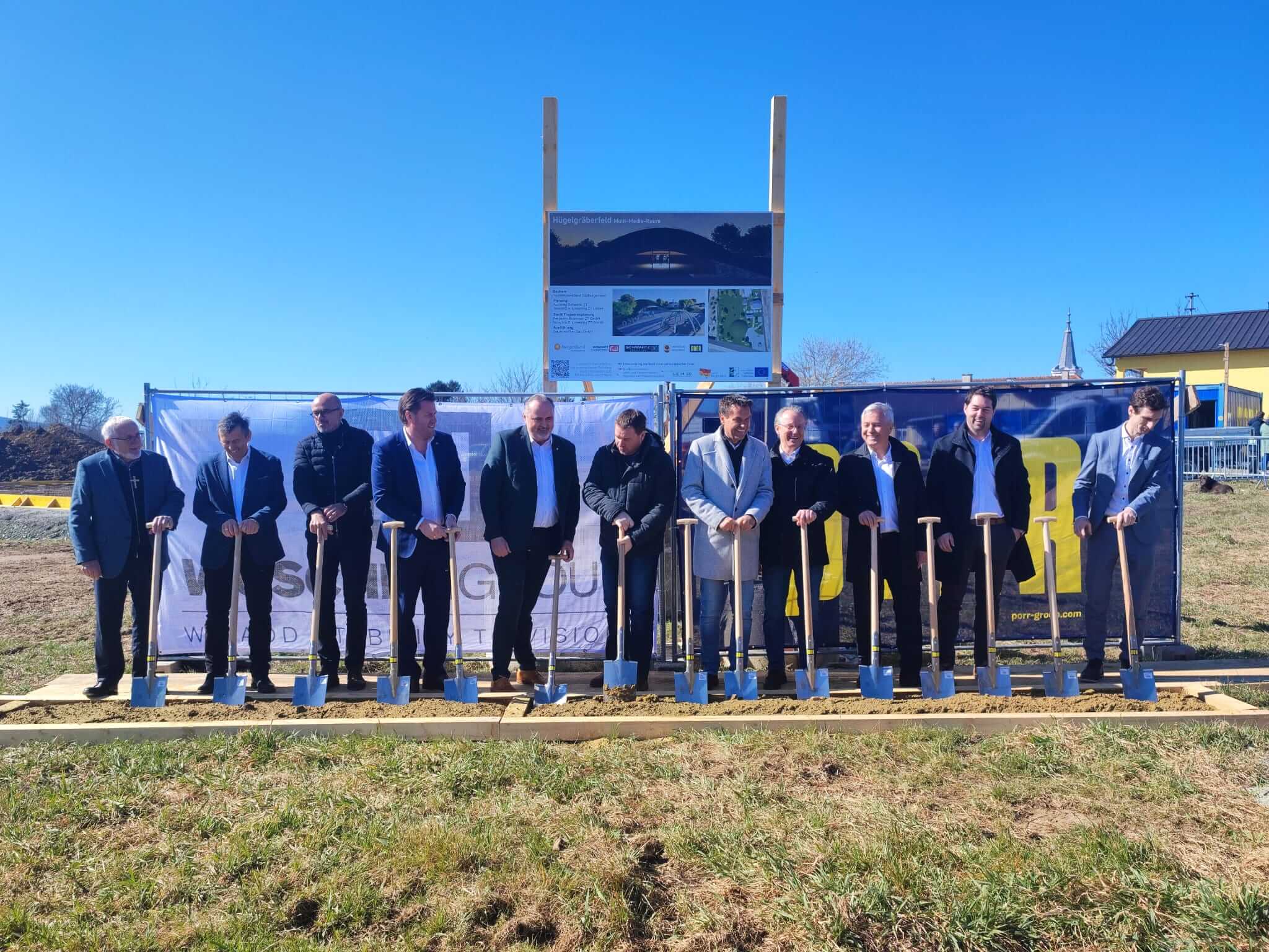 Gruppe von Menschen beim feierlichen Spatenstich auf einer Wiese vor blauem Himmel, Projektankündigung im Hintergrund.