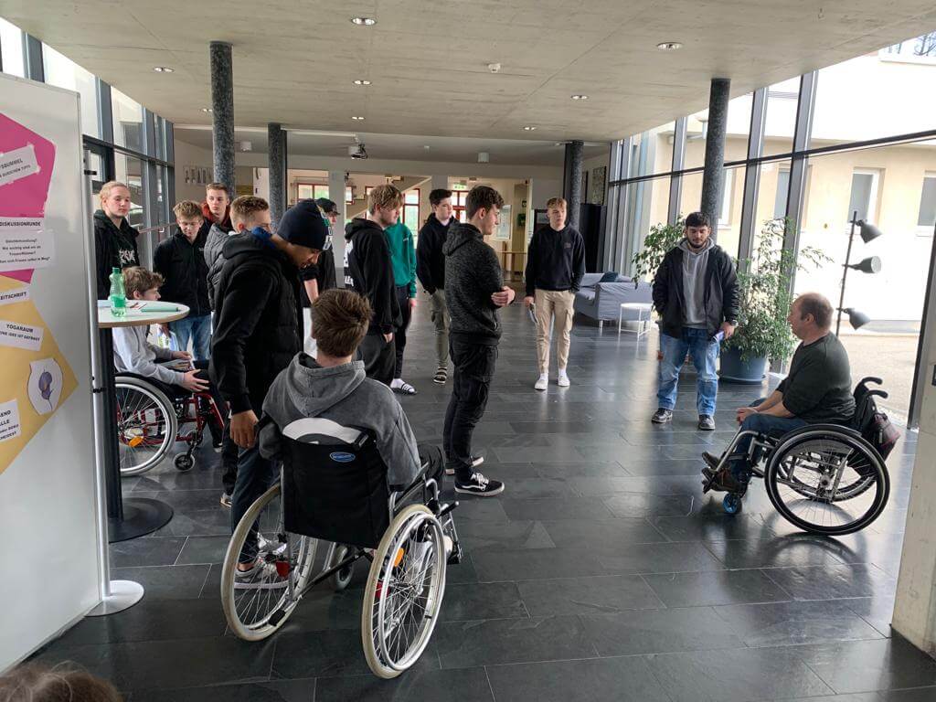 Personengruppe in einem modernen Flur, zwei Rollstuhlfahrer, Austausch und Gespräch über Barrierefreiheit.