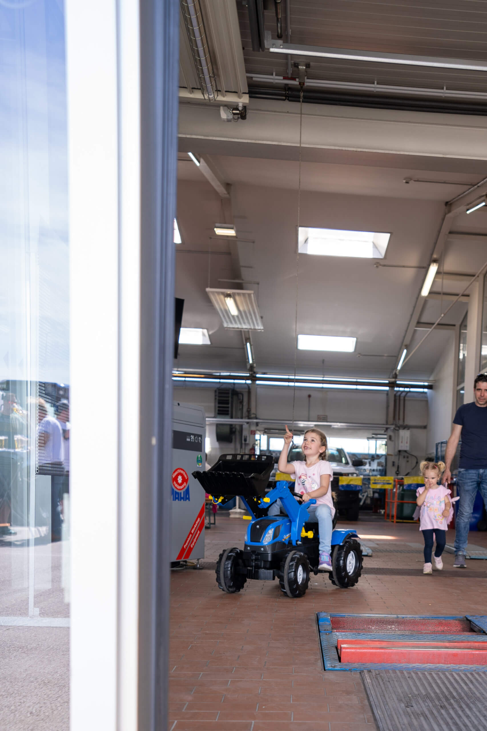 Kleines Mädchen auf blauem Spielzeug-Traktor in einer Werkstatt, begleitet von Erwachsenen und kleinem Kind im Hintergrund.