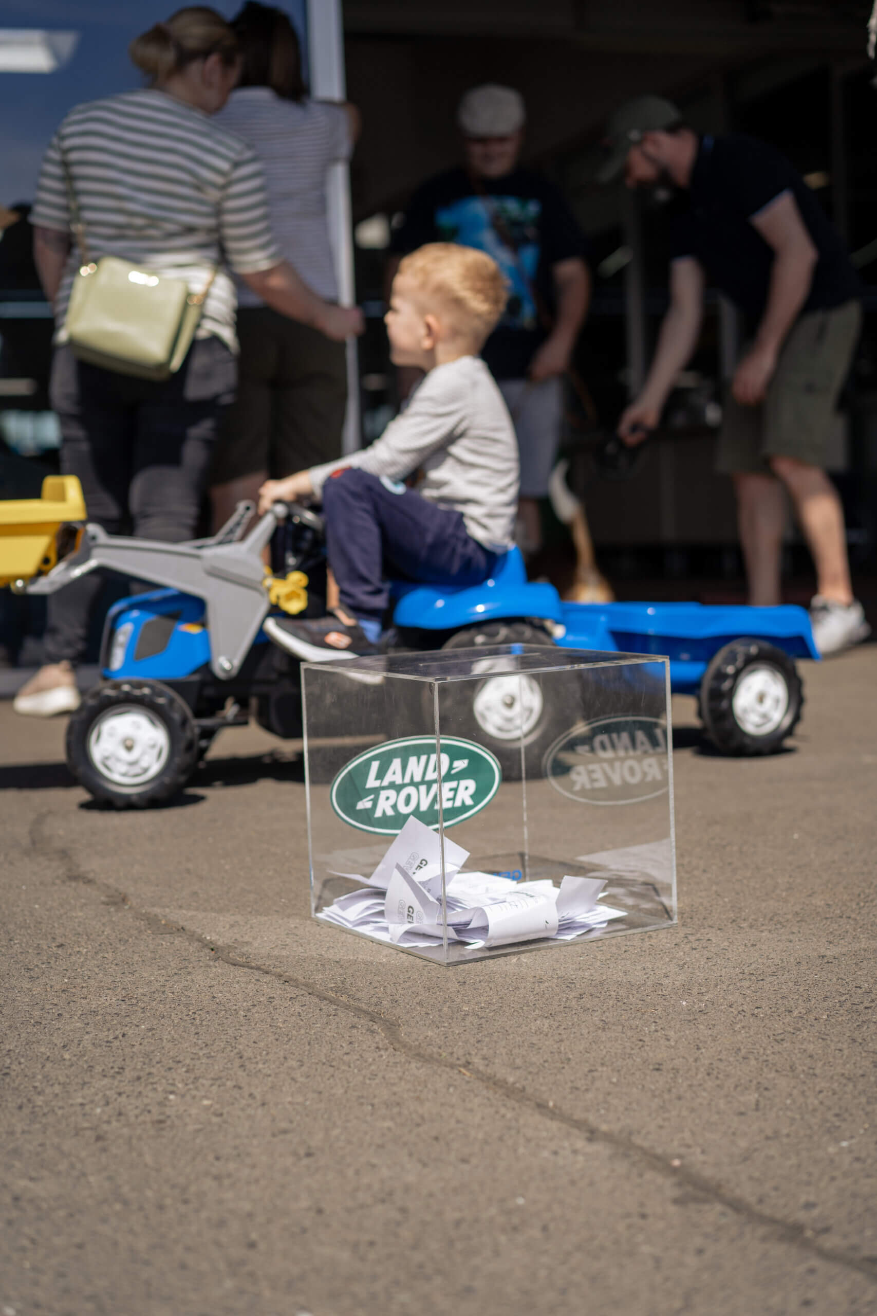 Kind auf Spielzeugtraktor neben Land Rover Werbebox, Familienevent im Freien, sonniger Tag.