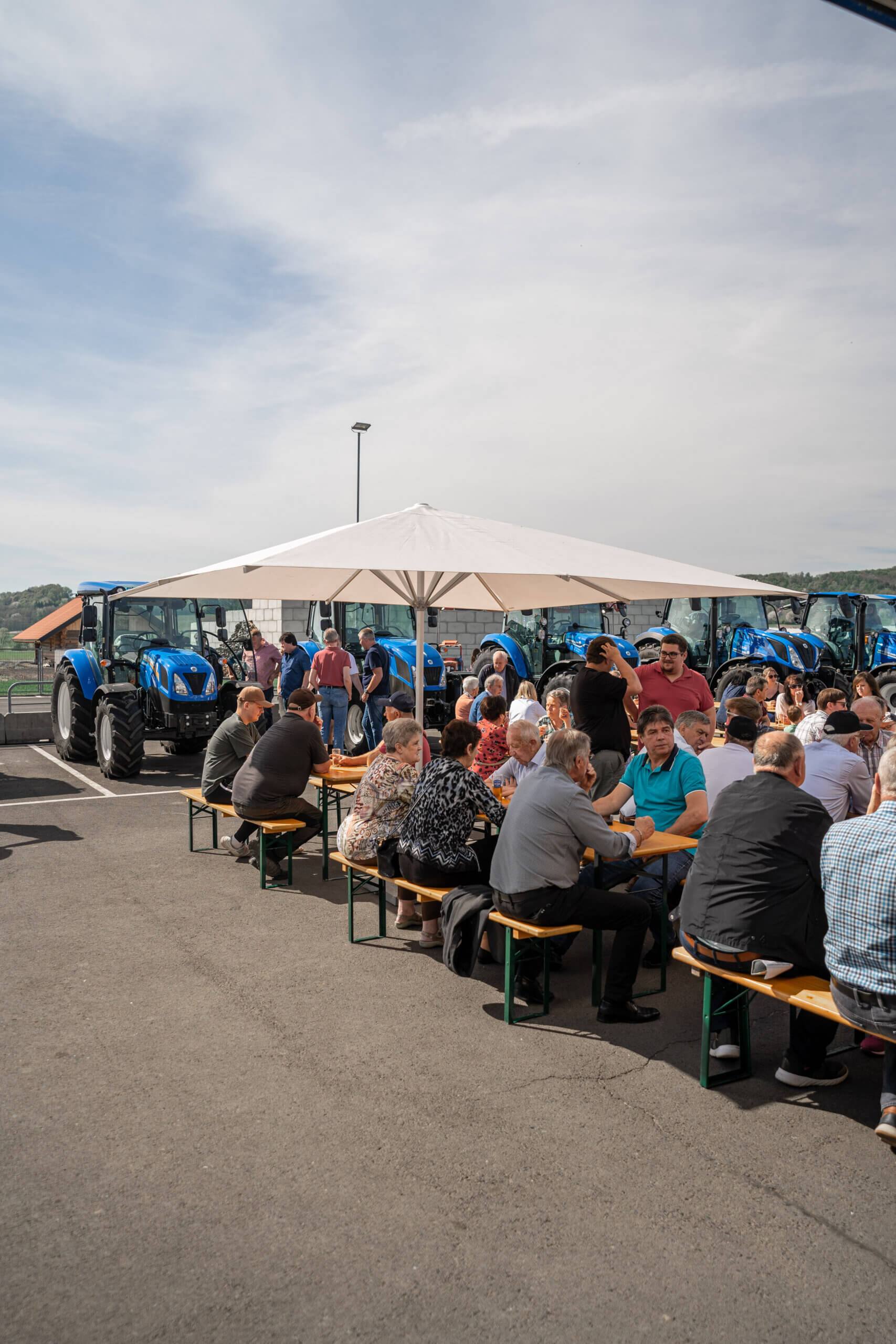 Gruppe von Menschen auf einem Hoffest mit Traktoren, unter einem Sonnenschirm an Biertischen sitzend.